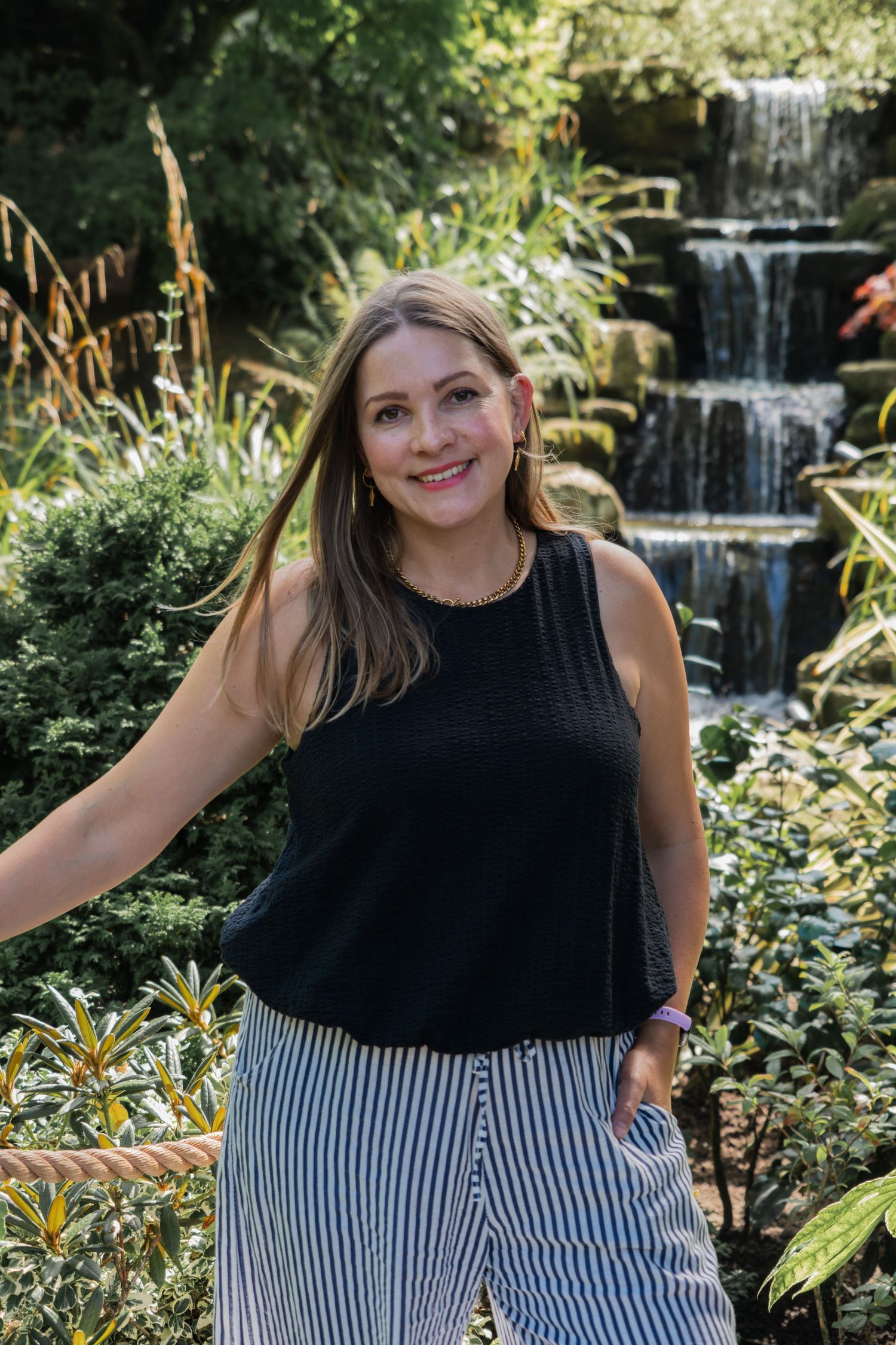 Woman in black top and striped skirt poses in front of garden waterfall in scenic outdoor photoshoot series.