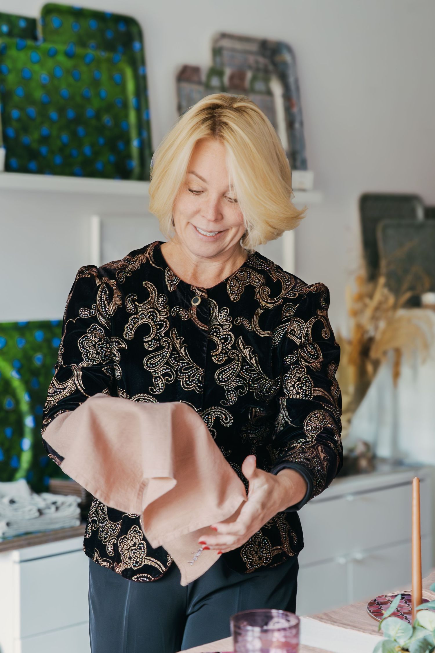 A person in a black embroidered top smiles while working with pink fabric in a bright interior space.