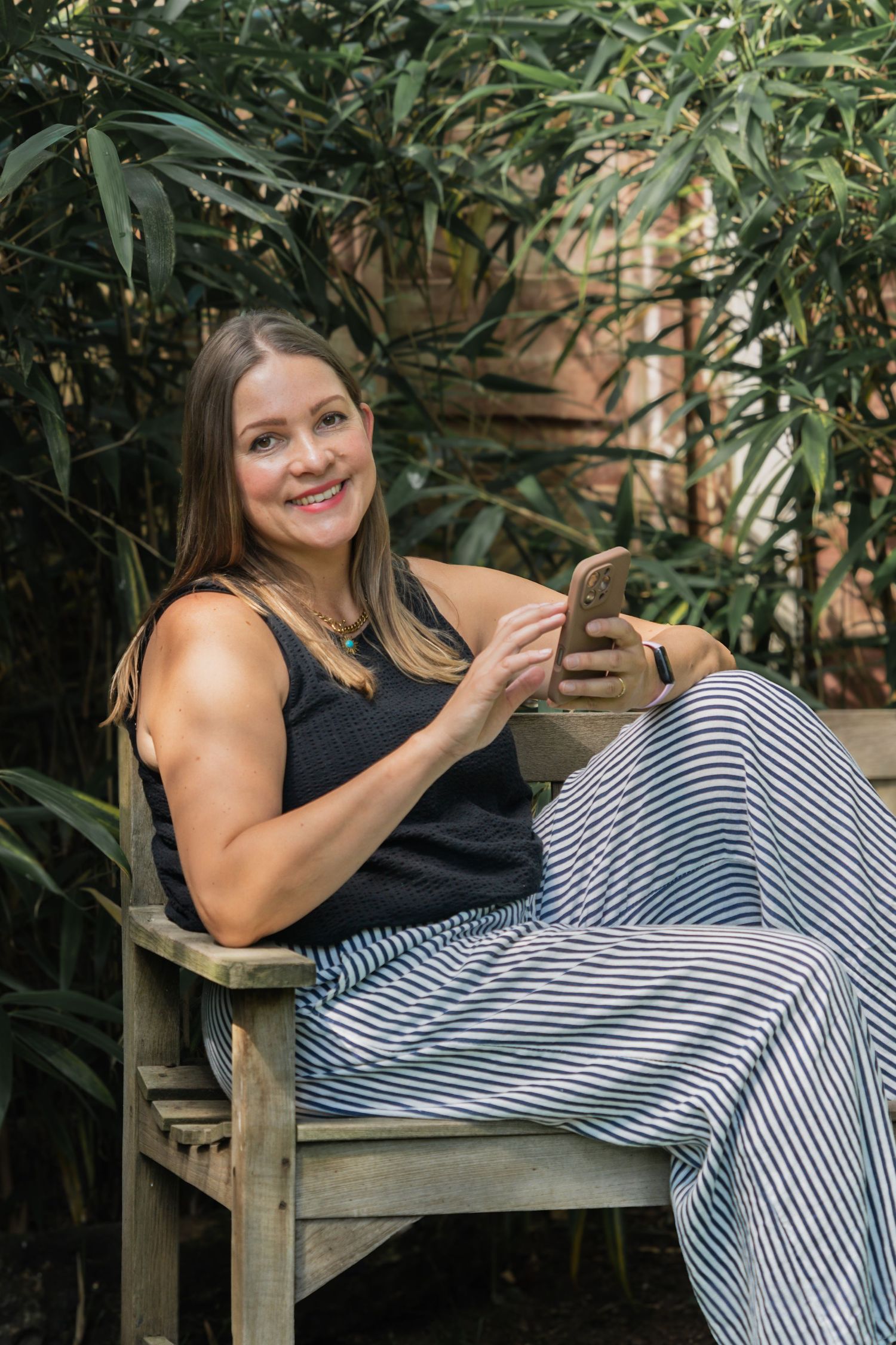 A person in a black top and striped pants relaxes on a wooden bench surrounded by lush greenery.