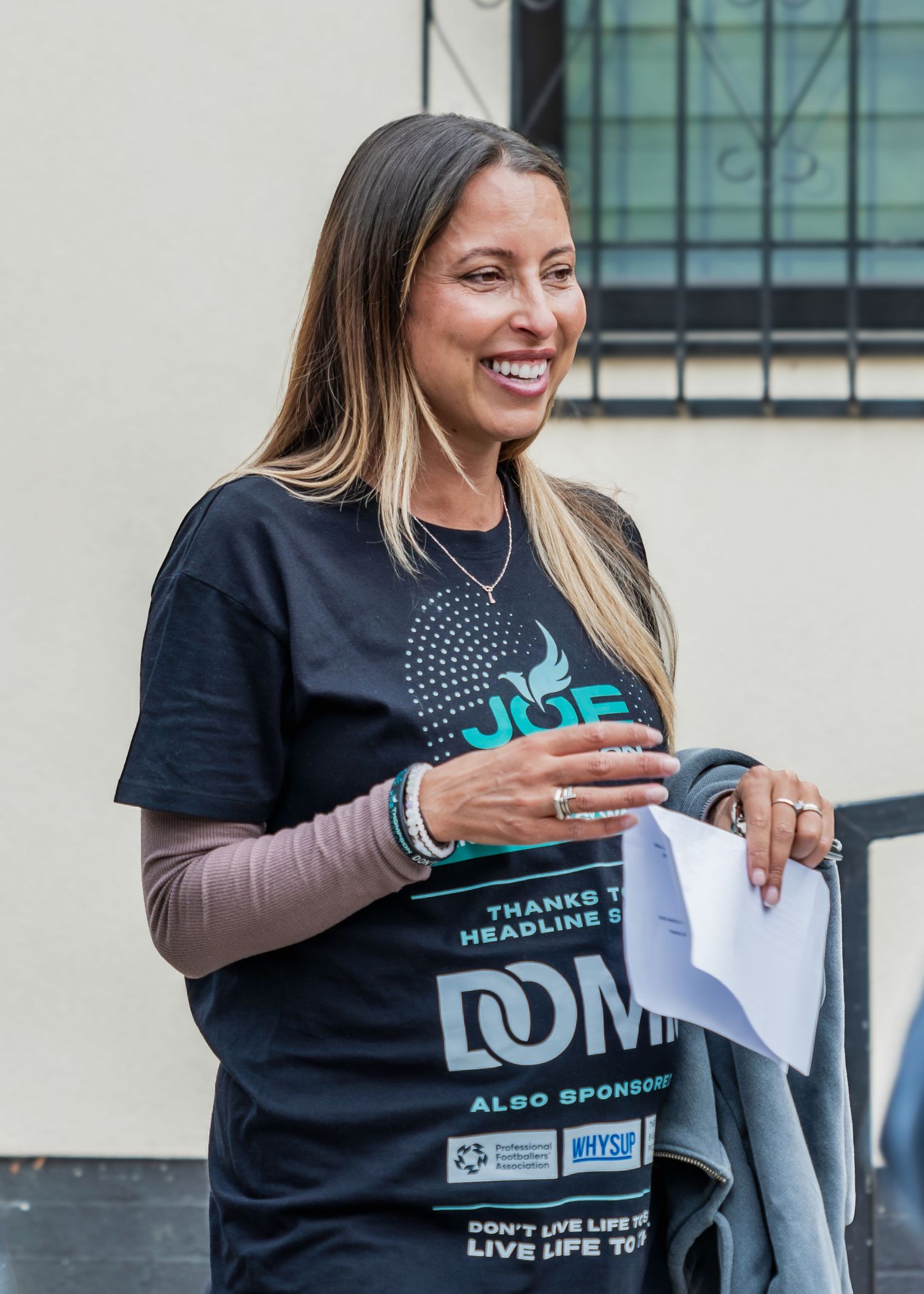 Smiling woman in a dark event t-shirt holding papers outdoors at a community gathering.