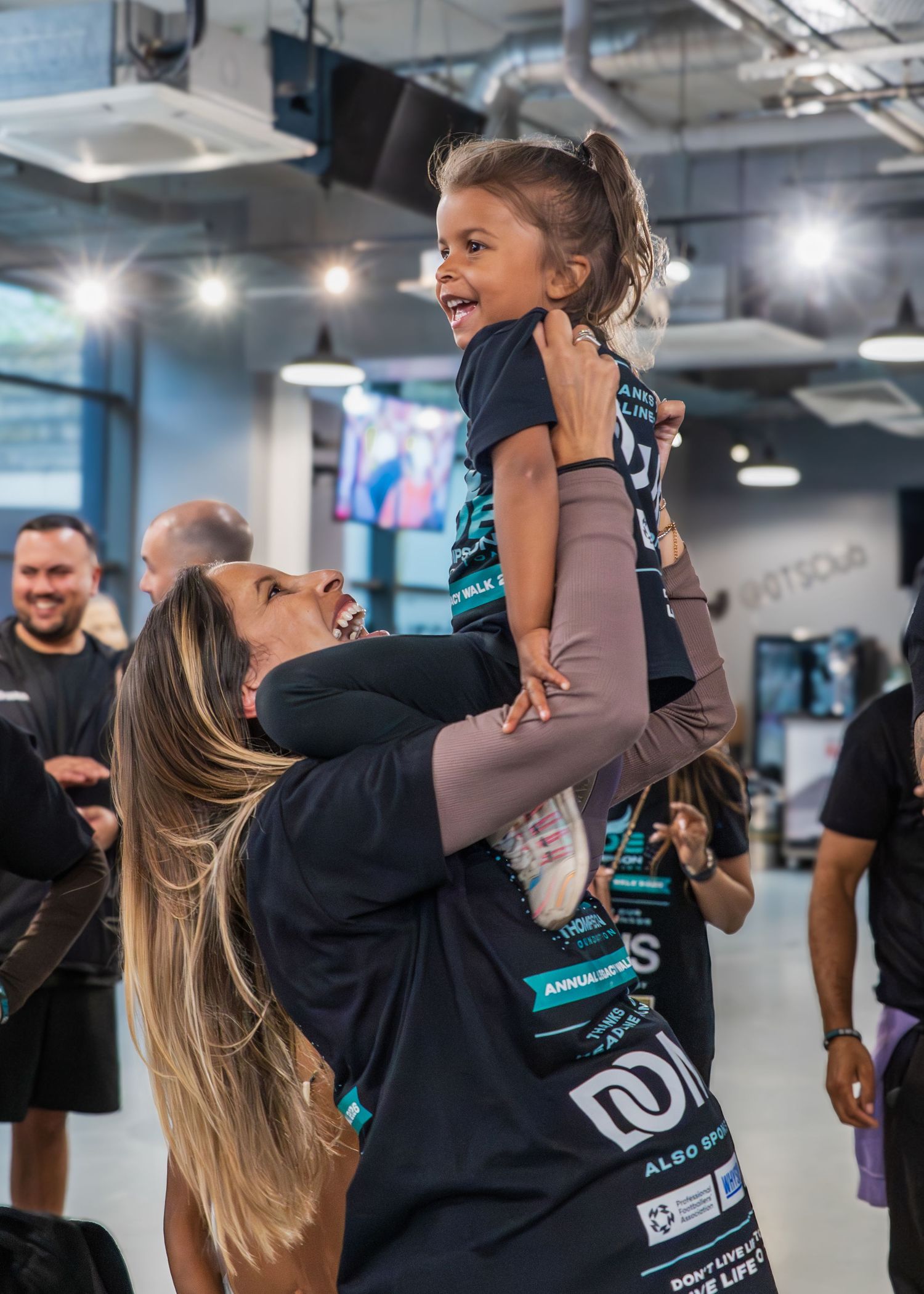 Woman joyfully lifting a smiling young girl in a gym, both wearing matching teal and black athletic shirts.