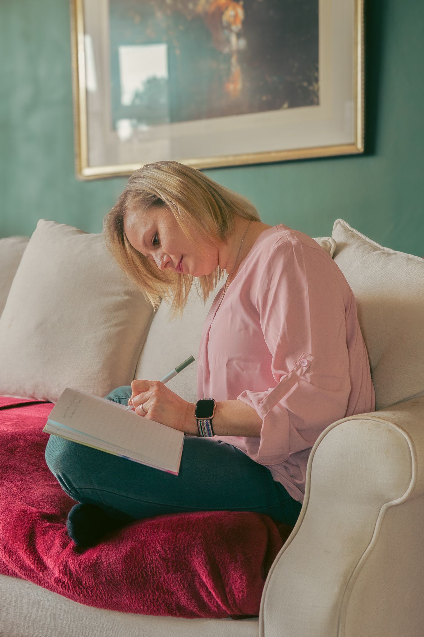 Someone wearing a pink shirt relaxes on a white couch with a red blanket while writing in a notebook.