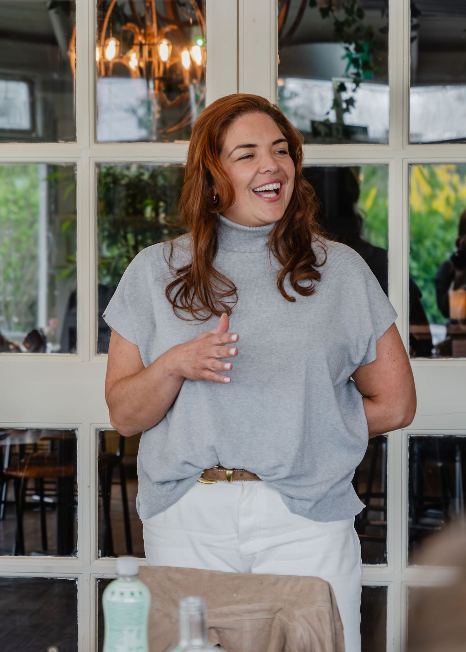 Smiling redhead woman in gray top and white skirt laughing warmly in a bright café setting.