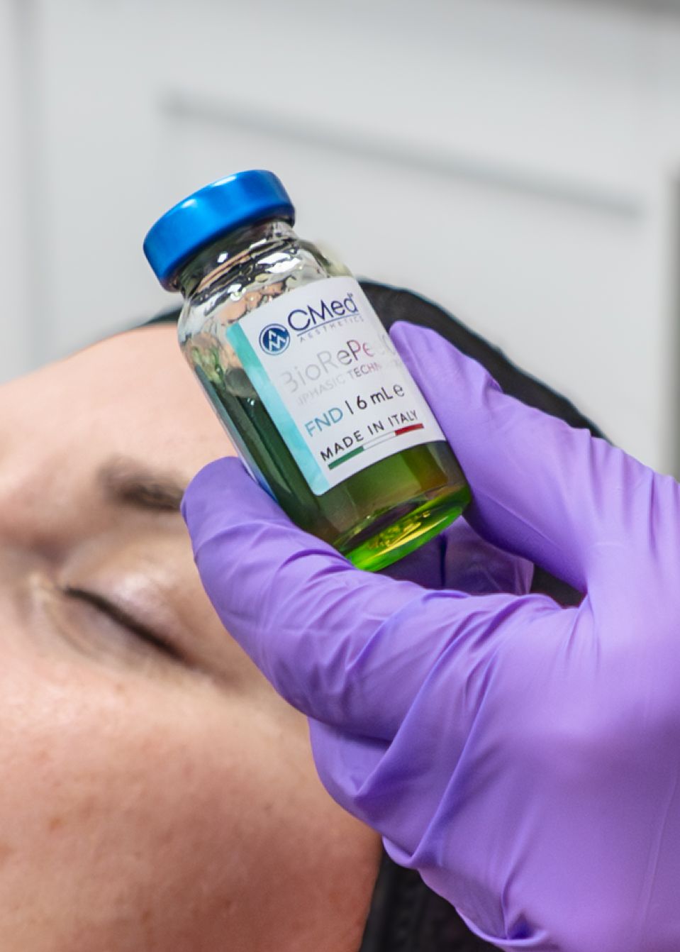 A gloved hand holds a CMed BioRePe PND 1.6mL vial made in Italy near a patient's face during a cosmetic treatment.