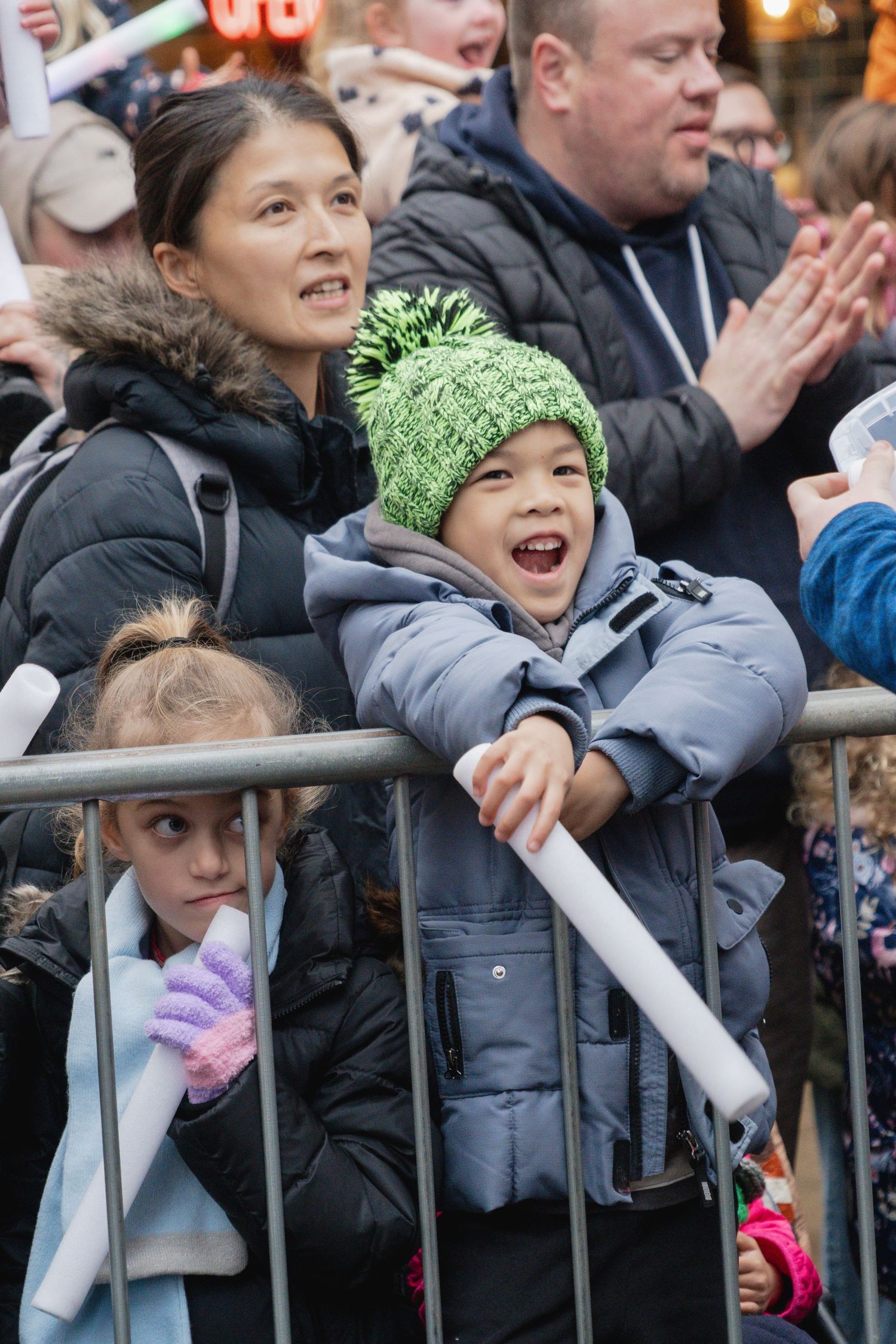Excited spectators lean over metal barriers at an outdoor event, with some wearing winter coats and knit hats.
