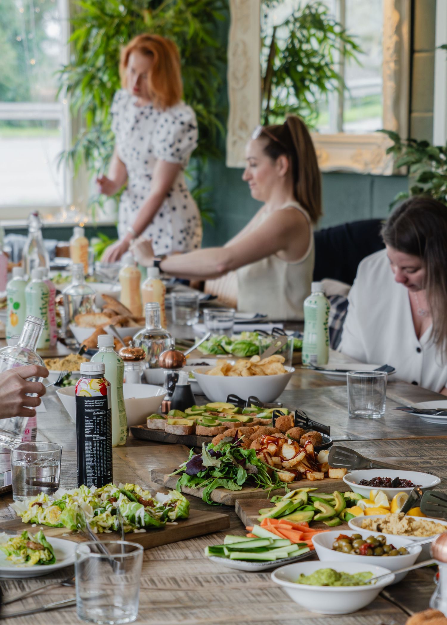 Women gathered around a table filled with fresh salads, dips, and healthy dishes at an indoor dining event.