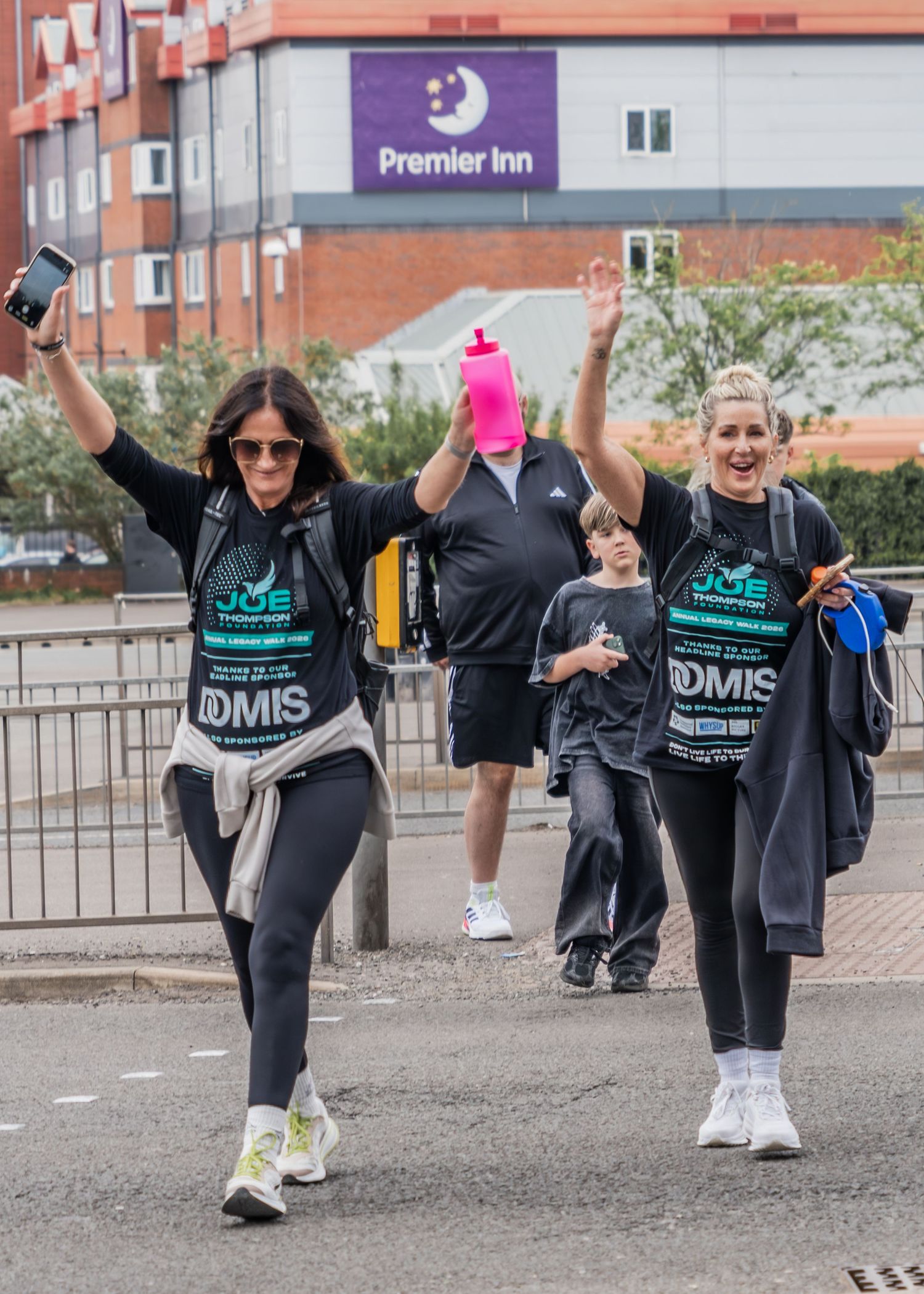 Two women in DOMIS charity walk bibs celebrate with raised arms near a Premier Inn hotel.