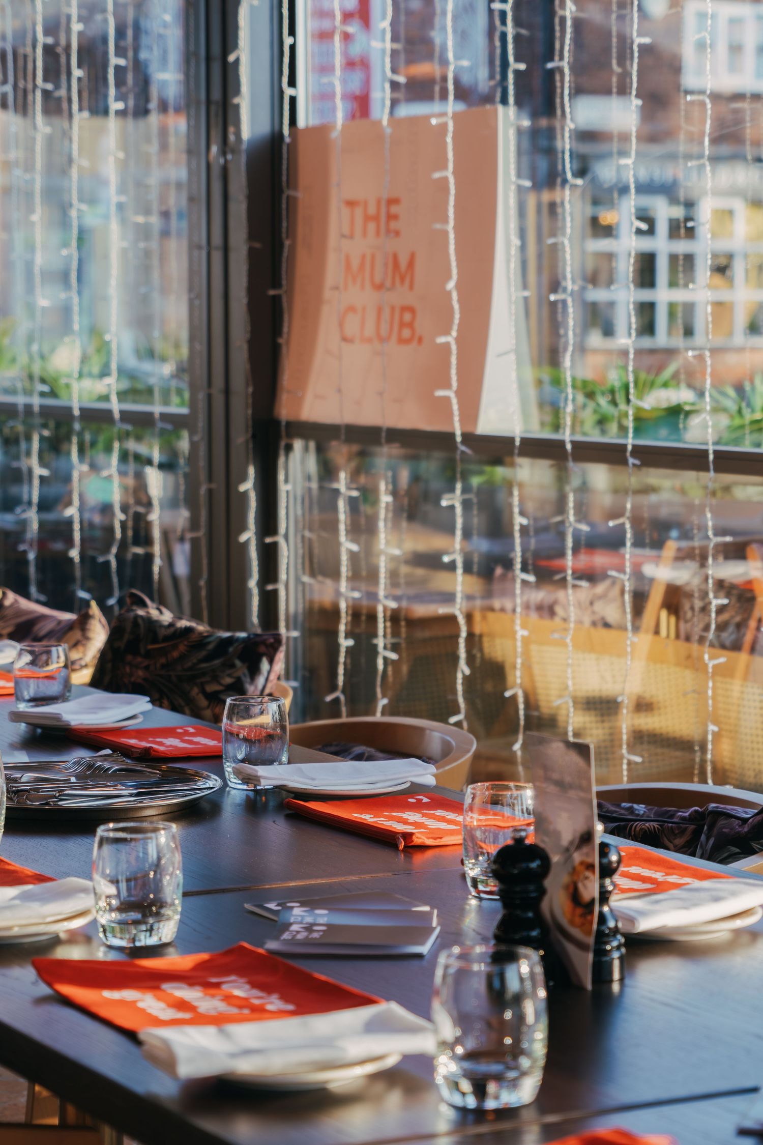 A modern restaurant dining table set with orange placemats and glassware in front of large windows.