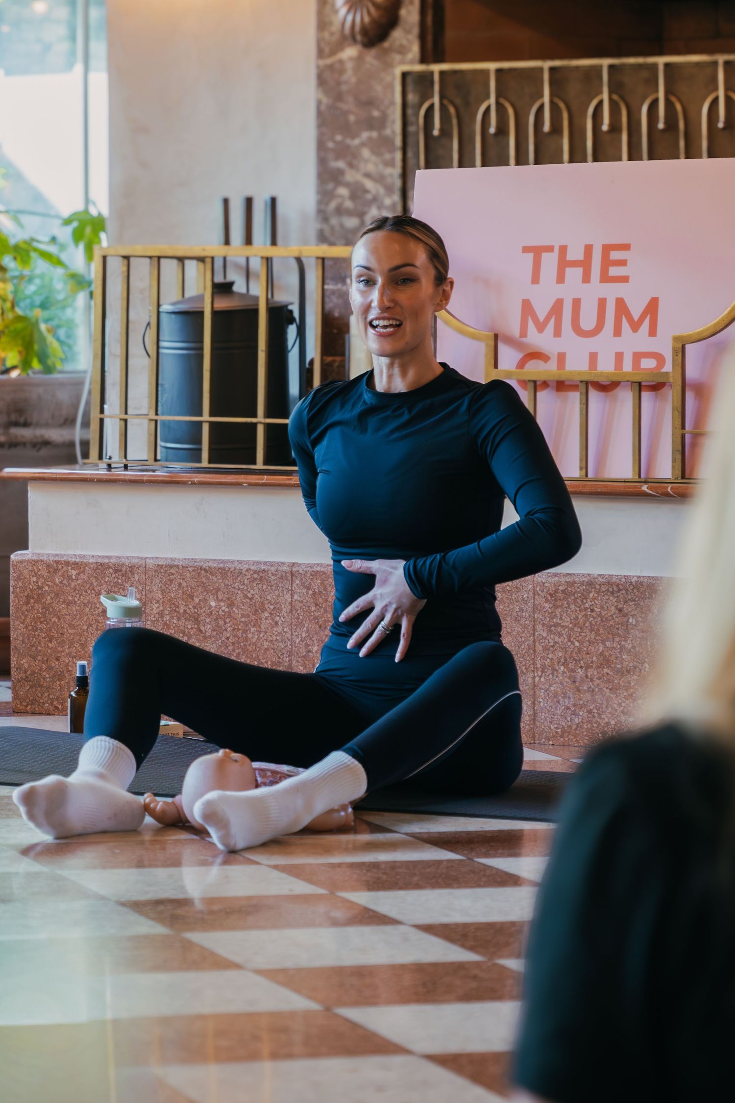 An instructor demonstrates a seated stretching position on the gym floor in a navy blue outfit.