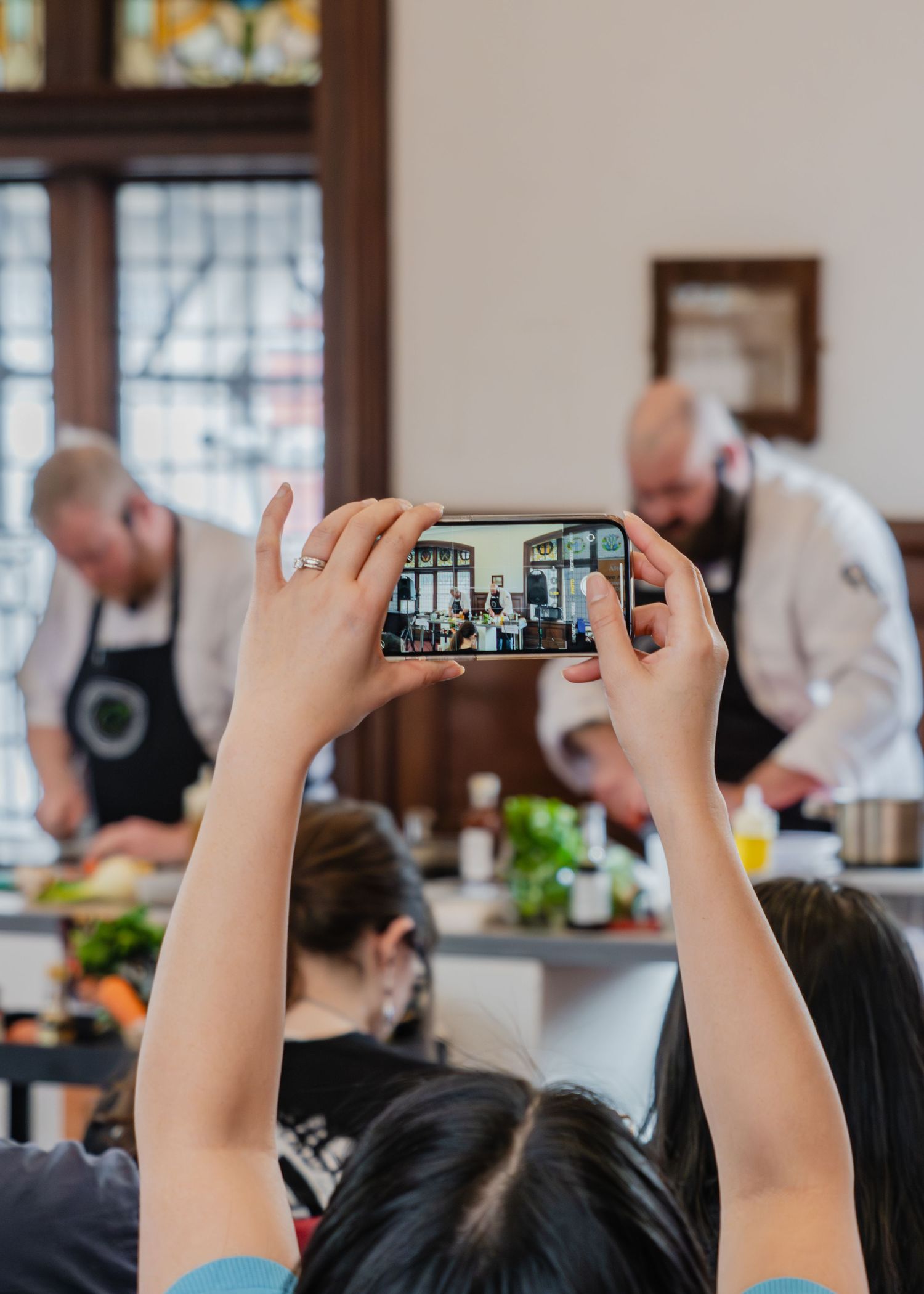Someone holds up a smartphone to photograph chefs working in a restaurant kitchen during a cooking class or demonstration.