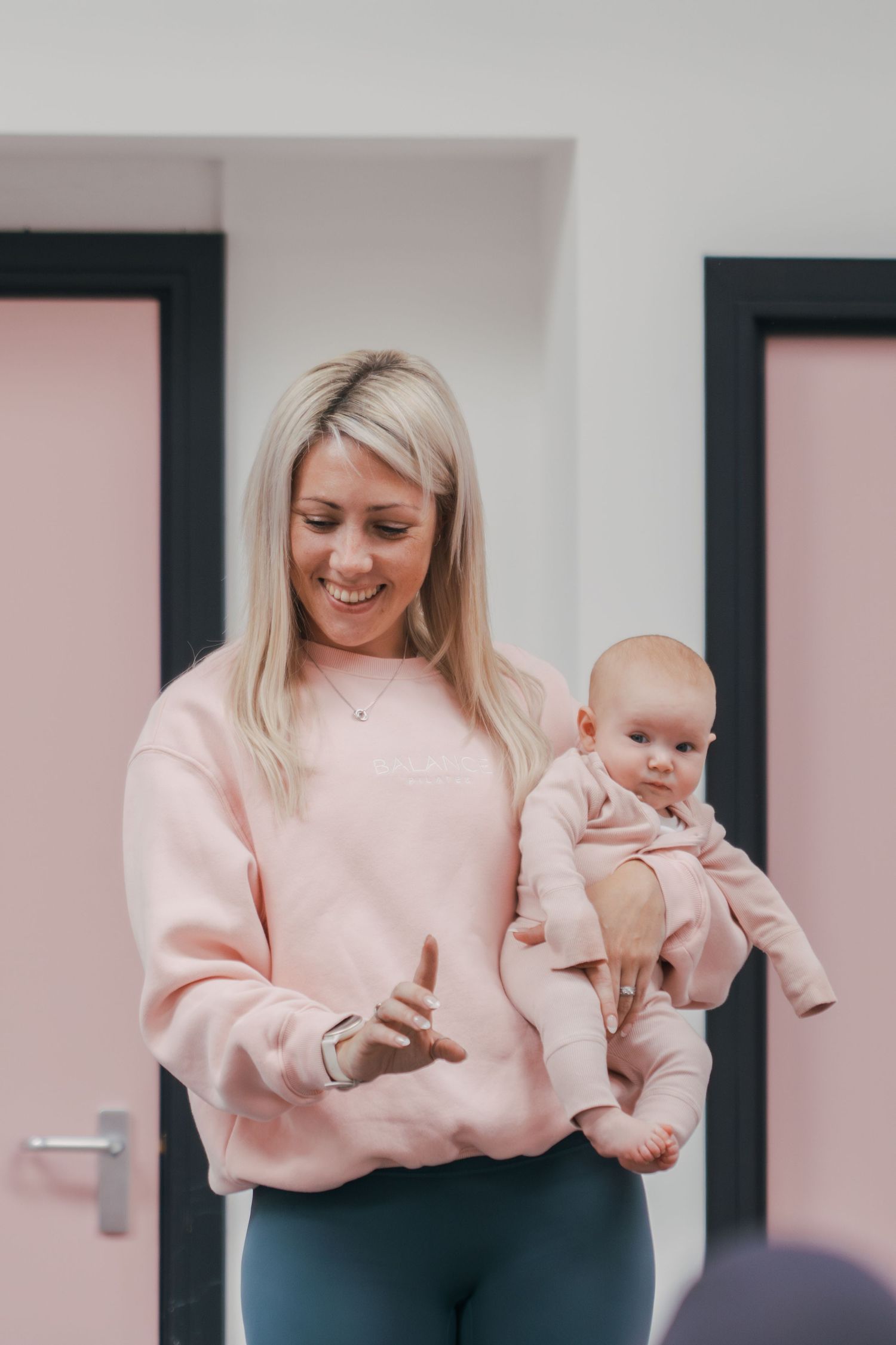 Parent in pink sweater holds and plays with baby in matching outfit against white interior wall.