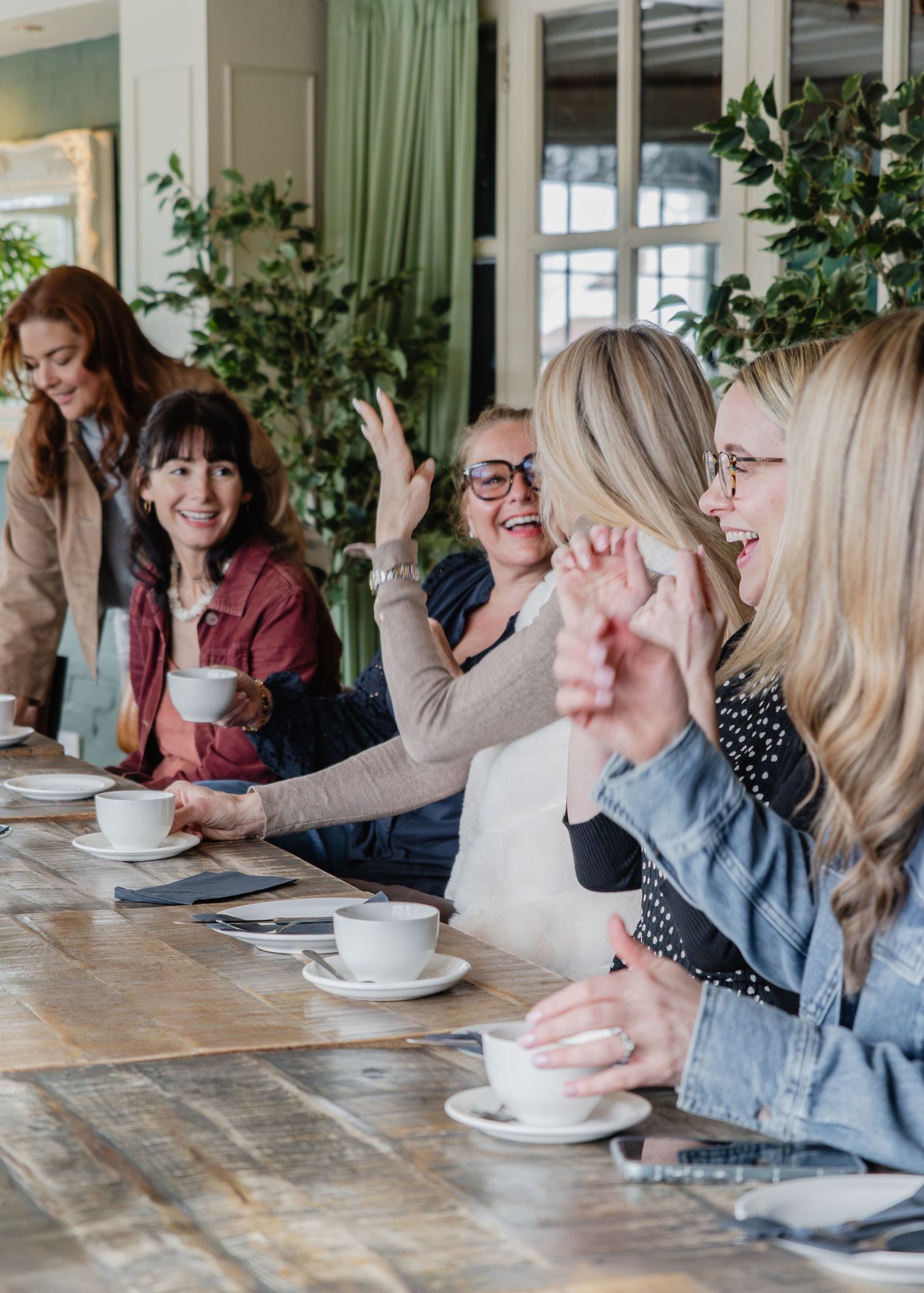 A group of women laughing and enjoying coffee together at a long wooden table in a bright, plant-filled cafe.