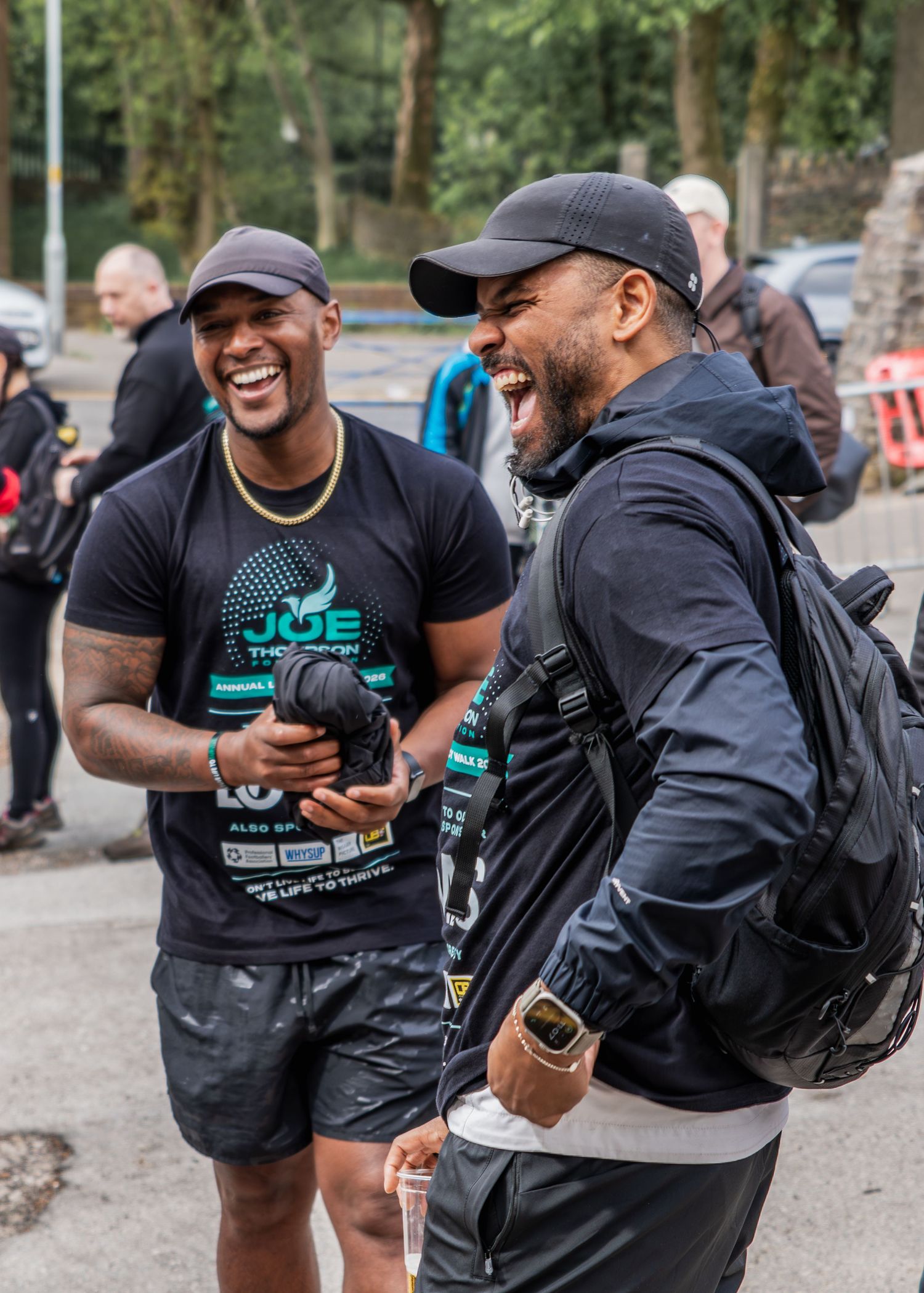 Two men in black Joe Thompson charity event shirts laugh together outdoors during a fundraising walk event.