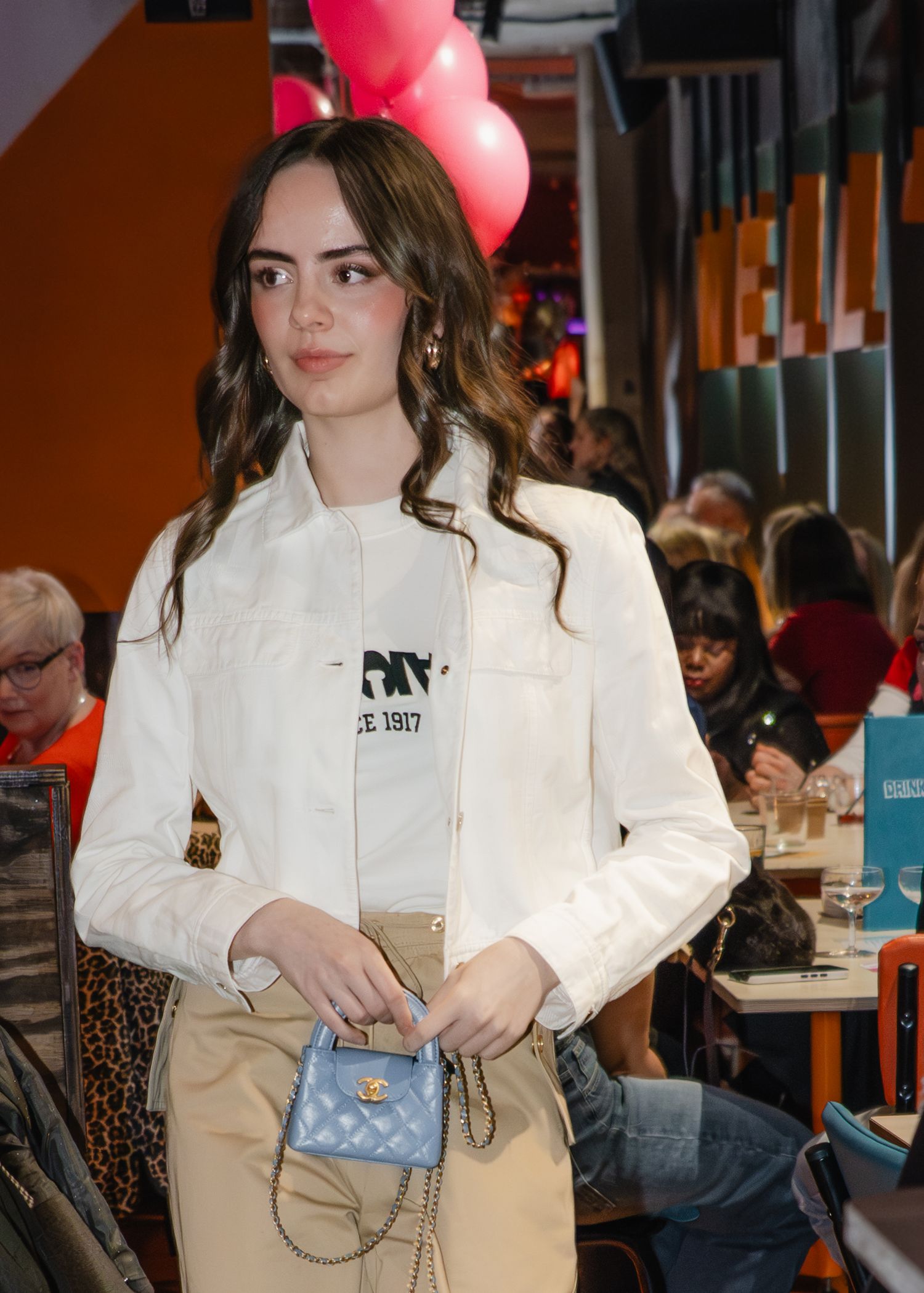 Young woman in white jacket holding a small blue quilted handbag at a restaurant event with pink balloons.