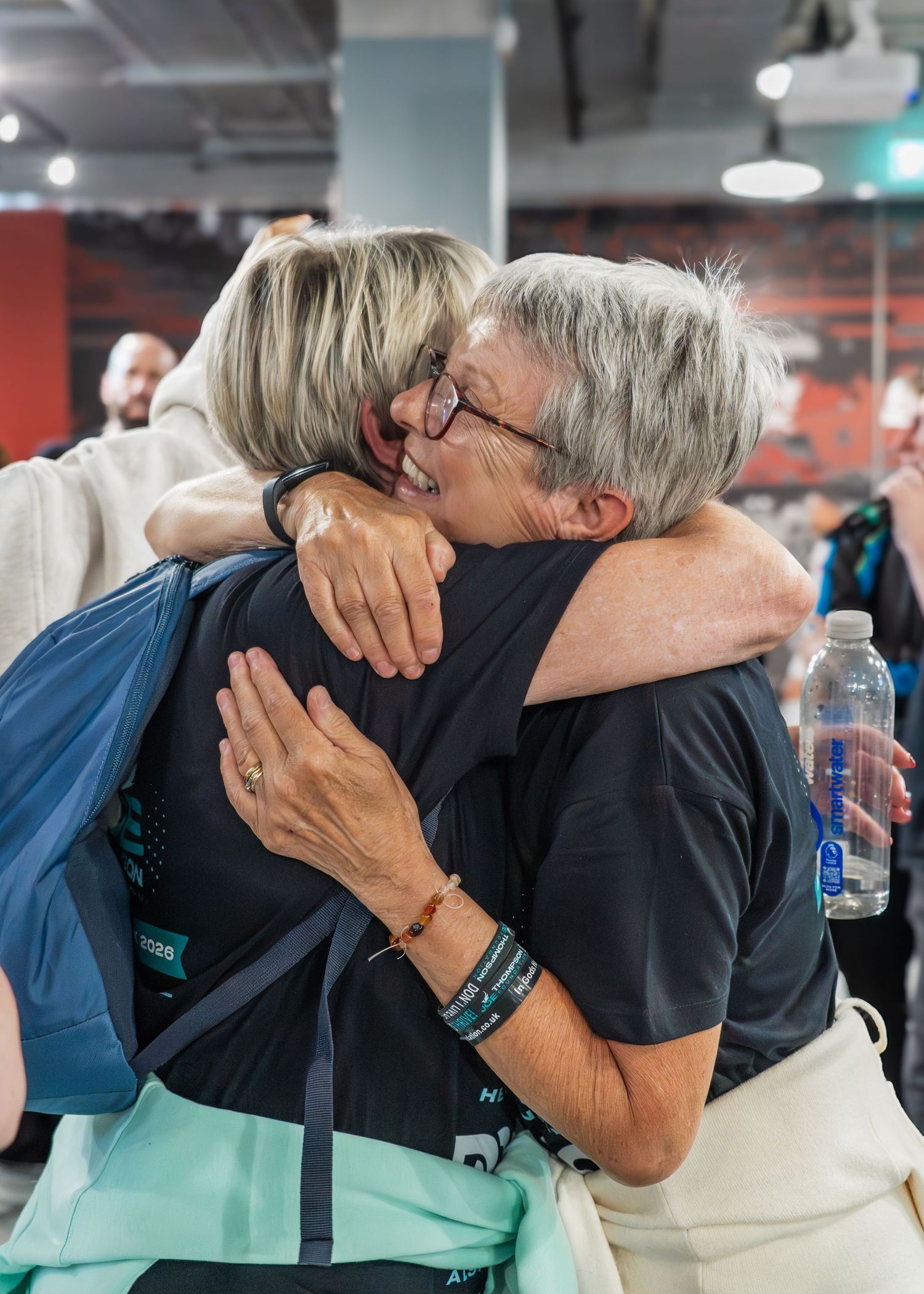 Two older women share a warm emotional hug at an indoor charity event finish line.