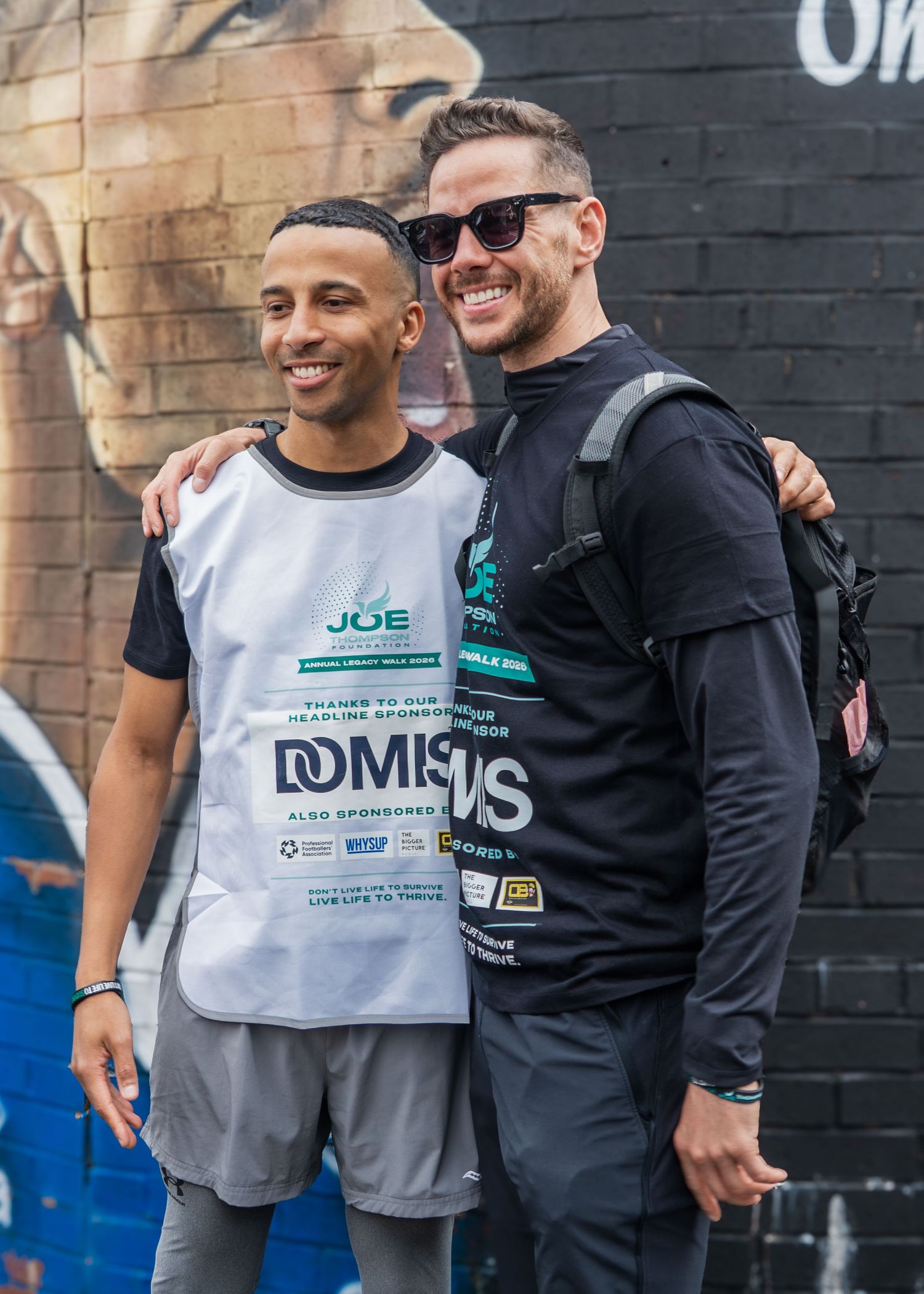 Two smiling men pose together at a charity running event near a street mural.