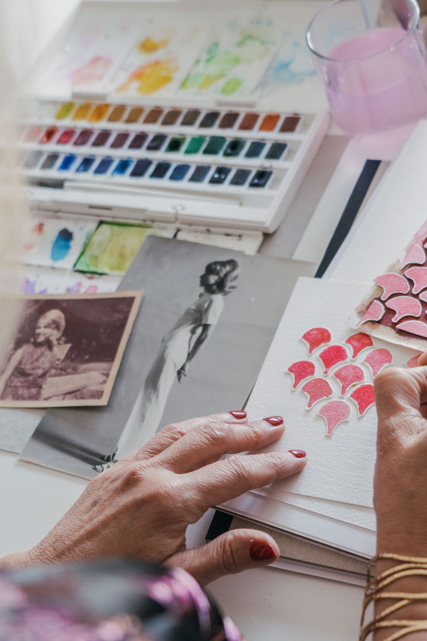 Close-up view of hands working on watercolor paintings with art supplies and reference photos spread across a white desk.