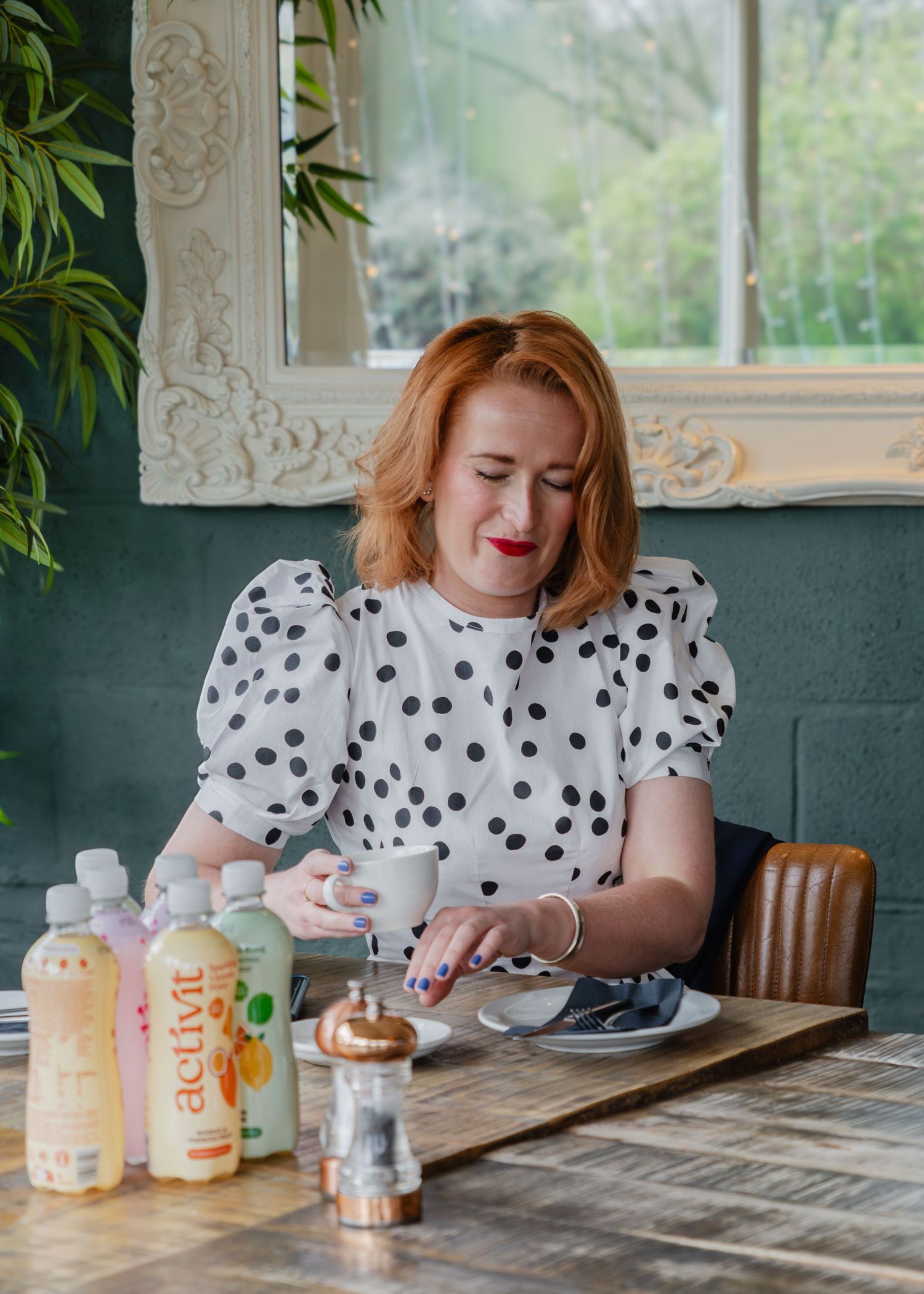 Red-haired woman in polka dot blouse sitting at cafe table with Activit drinks bottles, enjoying coffee.