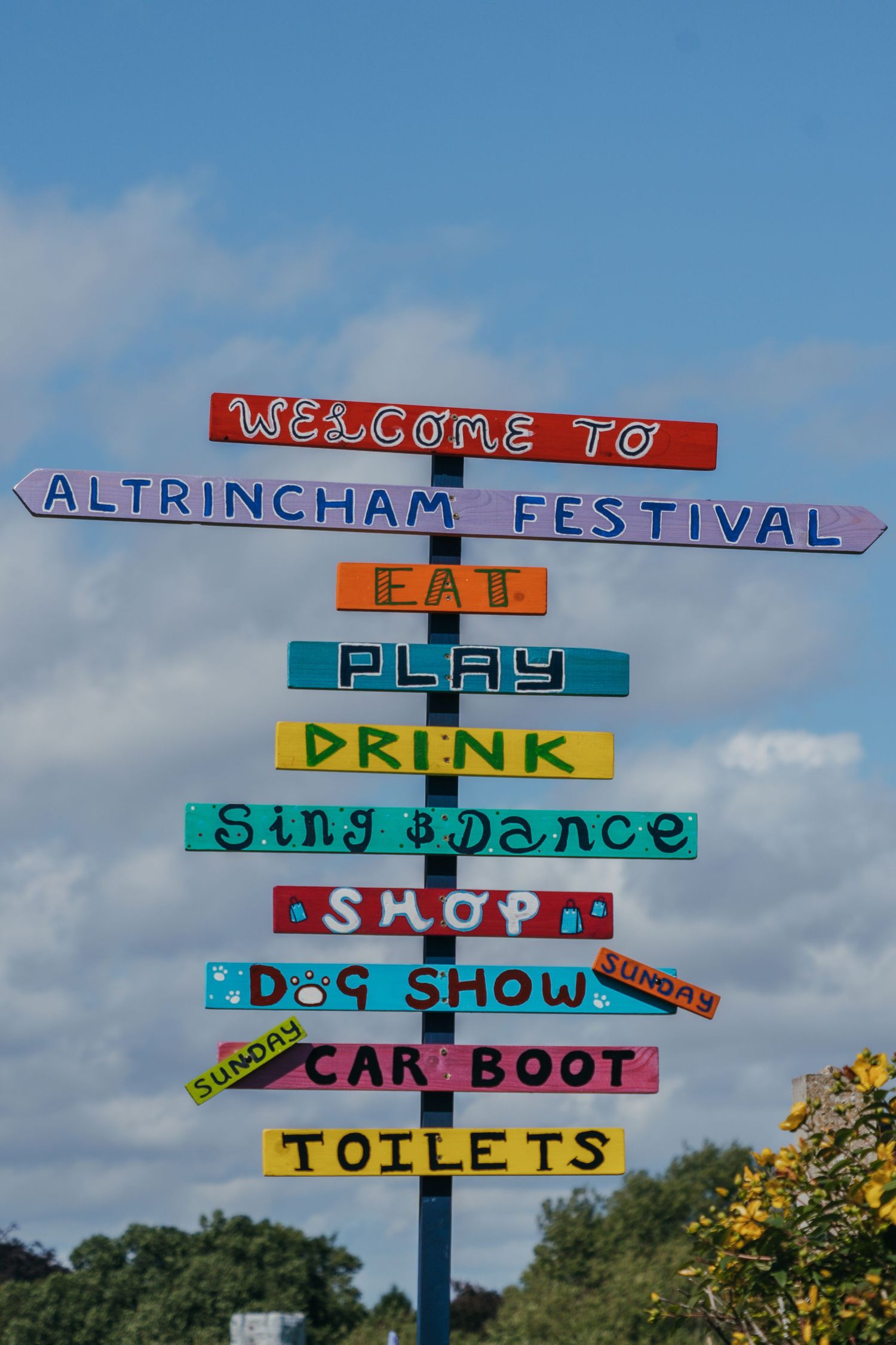 Colorful directional signs at an outdoor festival point to various attractions like toilets, car boot, and food stalls against blue sky.