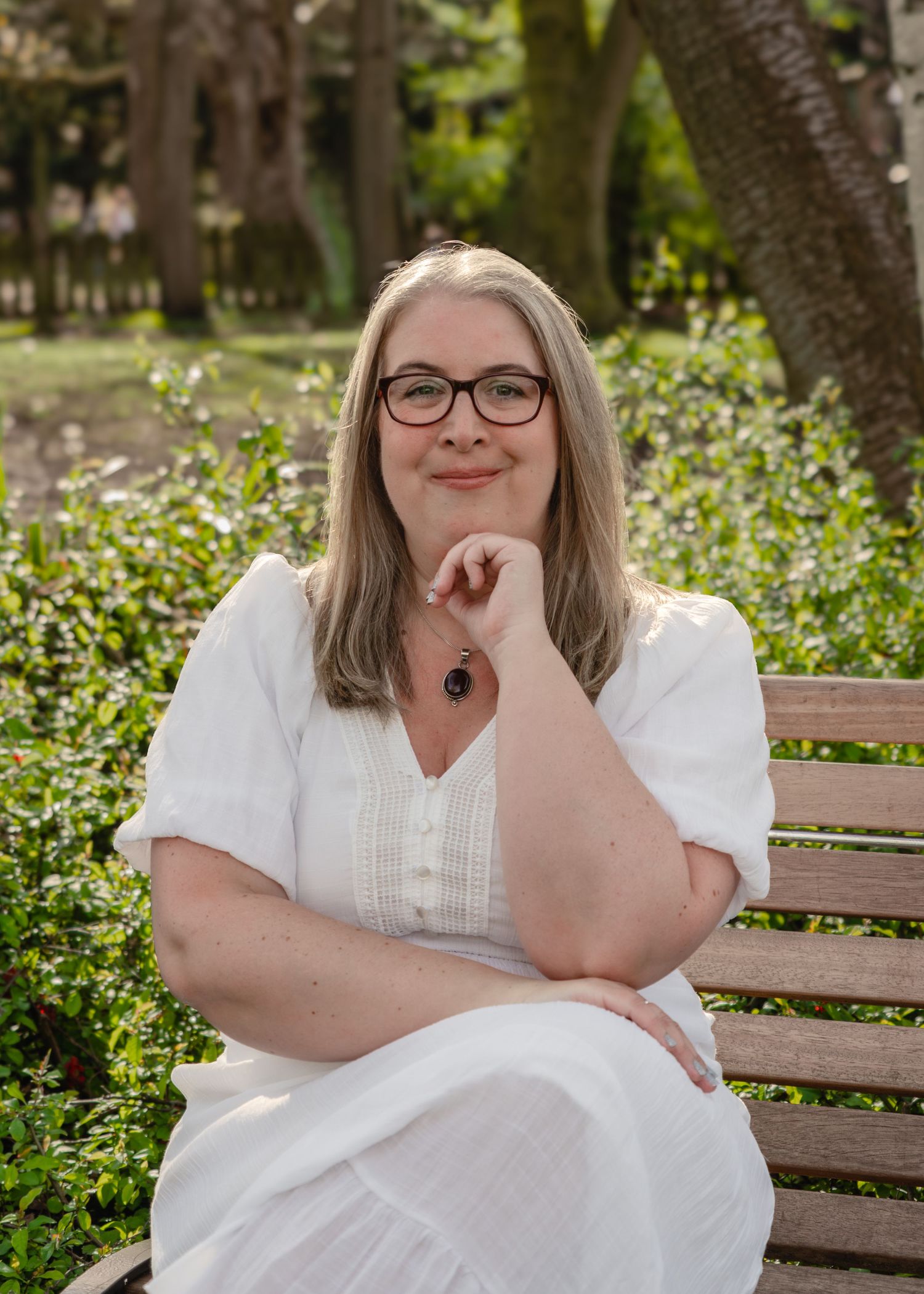 Woman in white dress sitting on bench in lush green park, smiling with glasses and long gray hair.