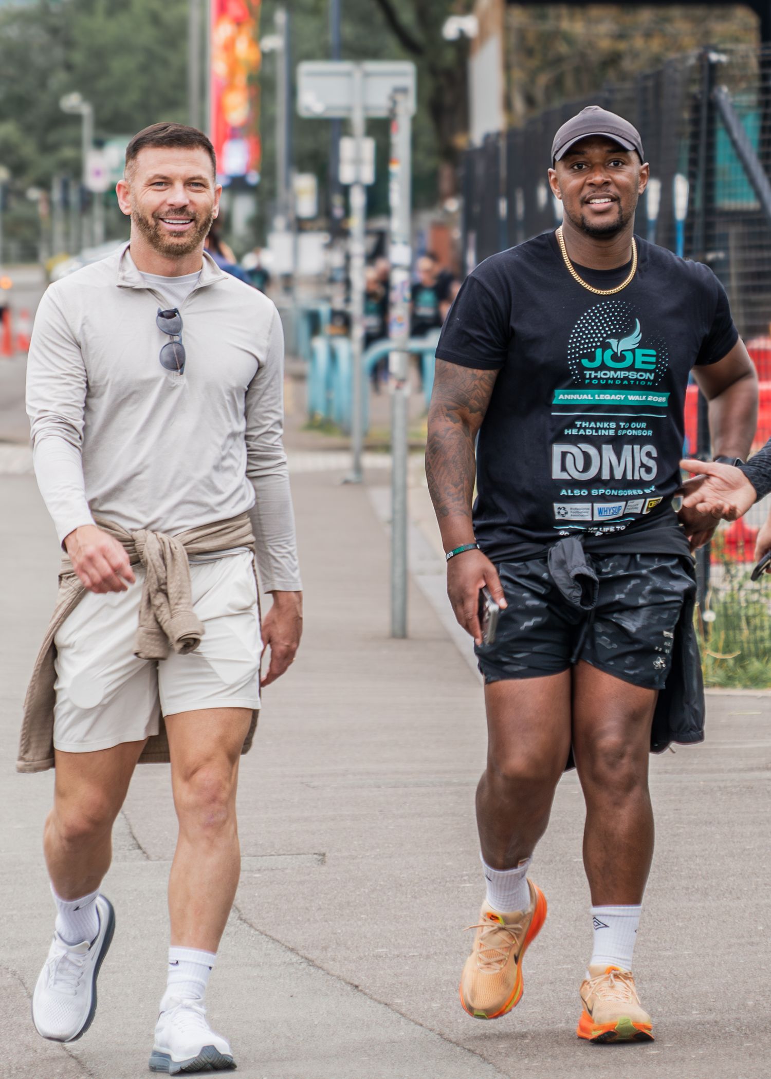 Two athletic men walking outdoors, one in gray outfit with sunglasses, other in black shirt and shorts with orange sneakers.