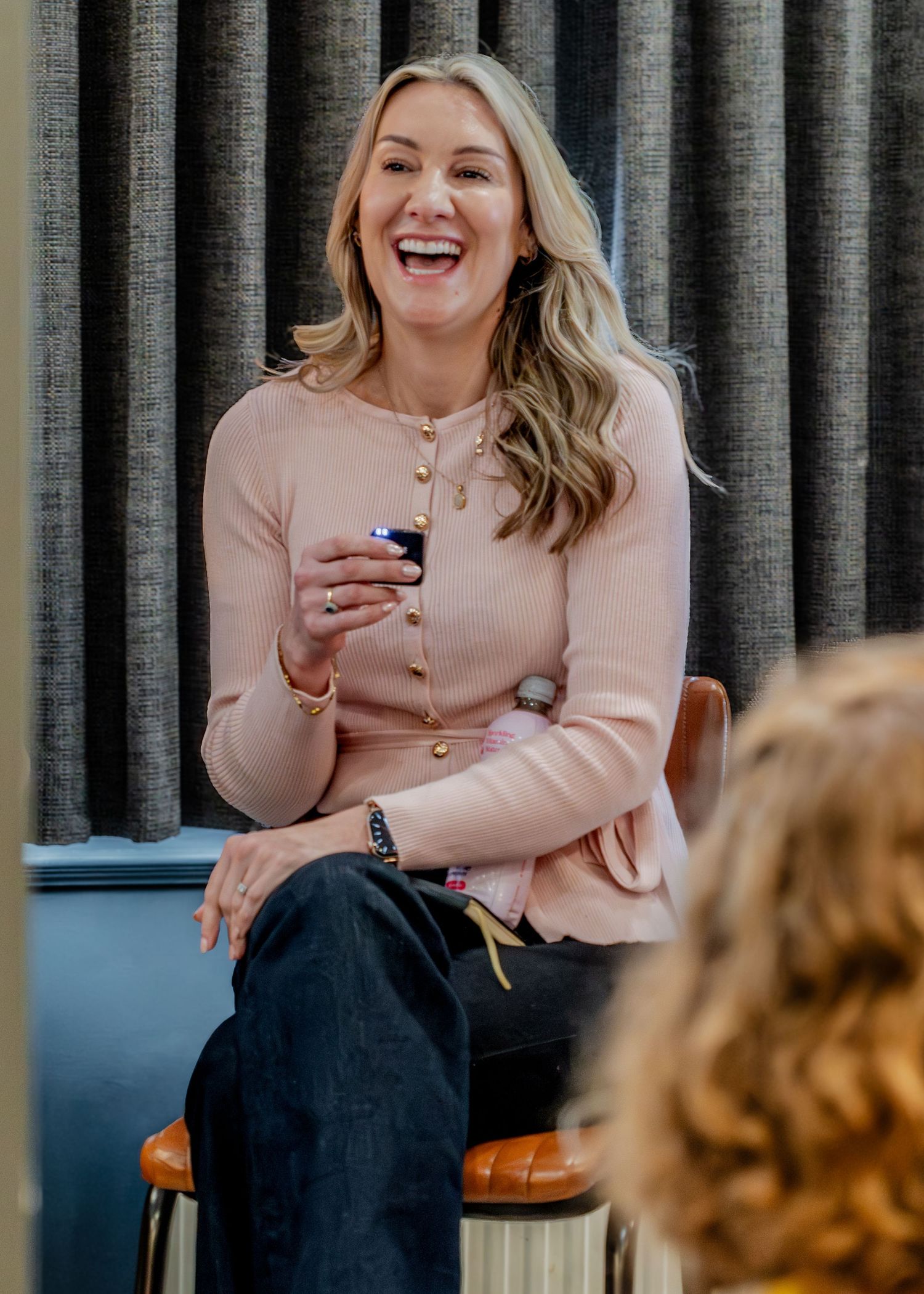 Smiling blonde woman in a pink cardigan laughing while seated during a presentation or panel discussion.