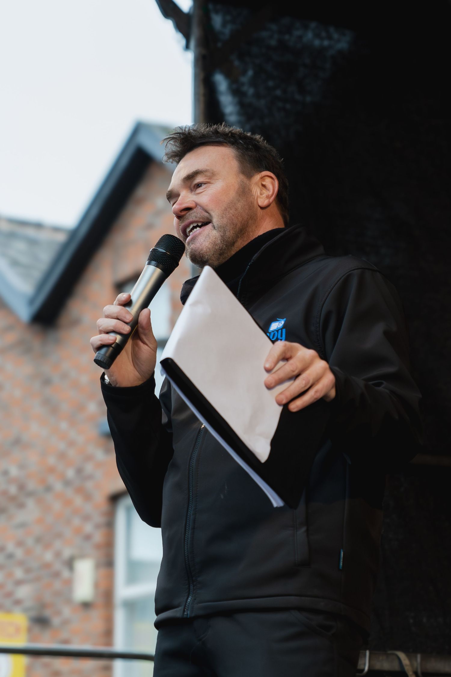 Person in black jacket laughing while holding papers during outdoor presentation.