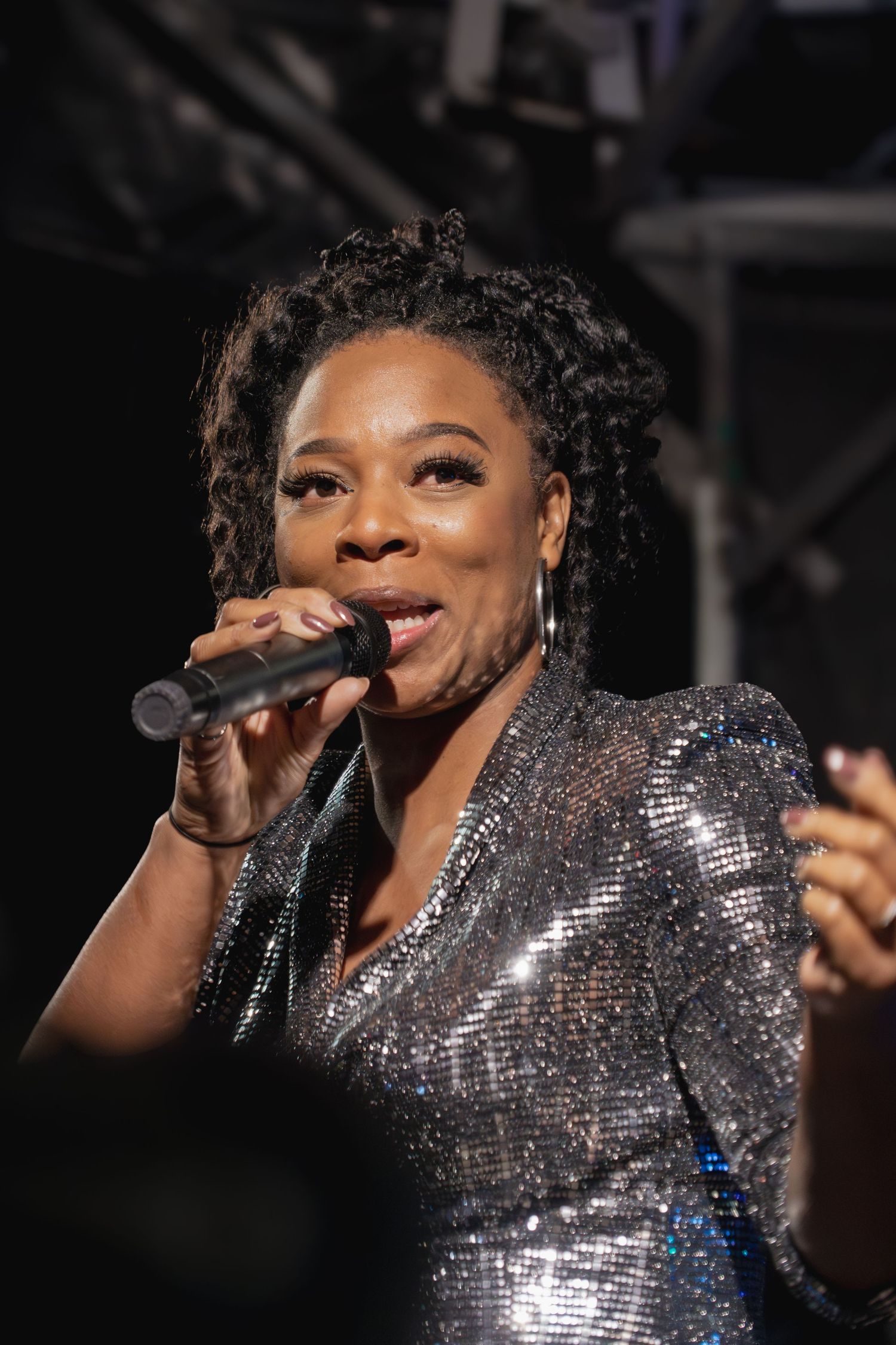 A performer in a sparkling silver dress sings passionately into a microphone during a live concert performance.