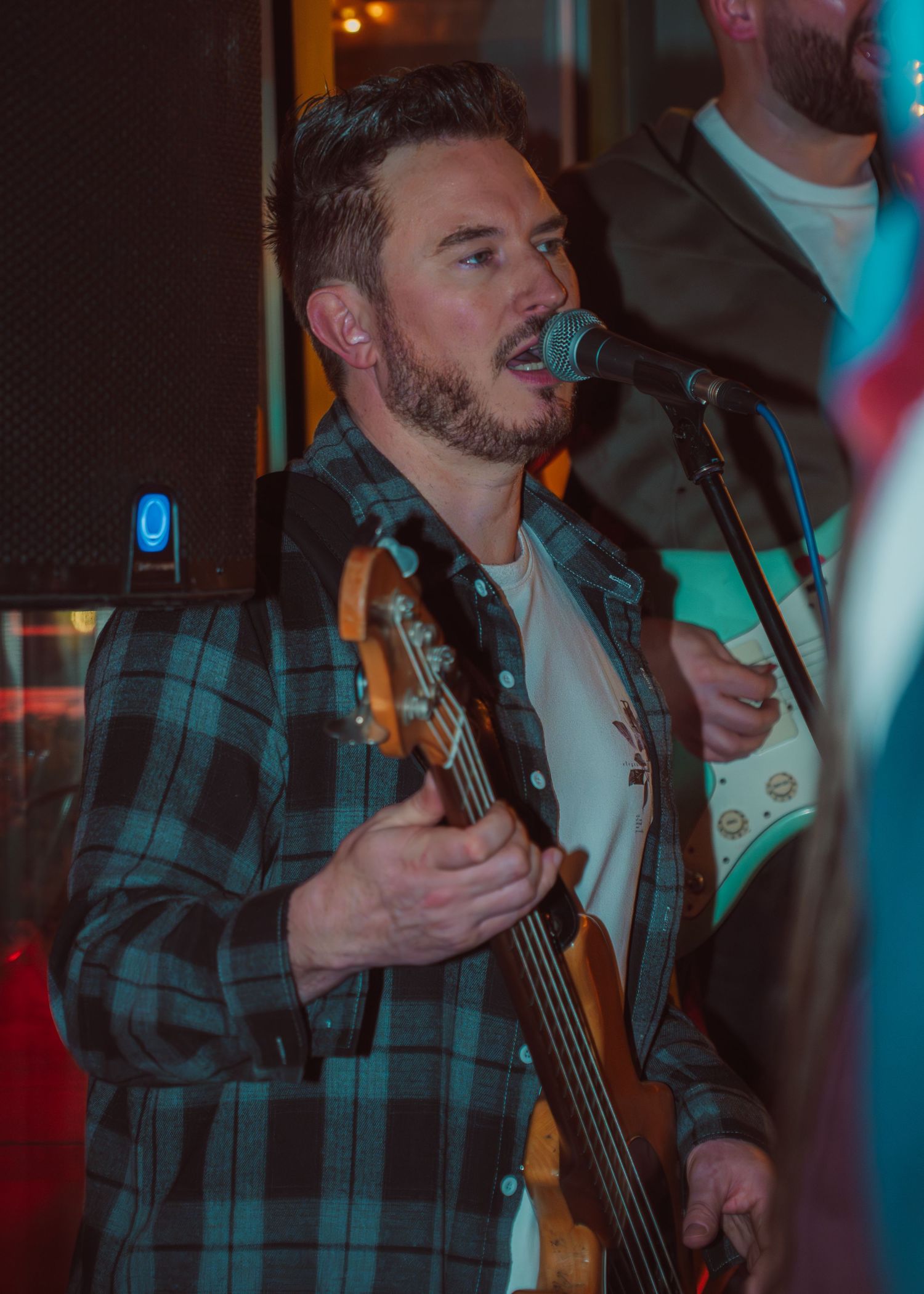 A musician performs with passion on stage in a plaid shirt under blue and pink lighting during a live show.