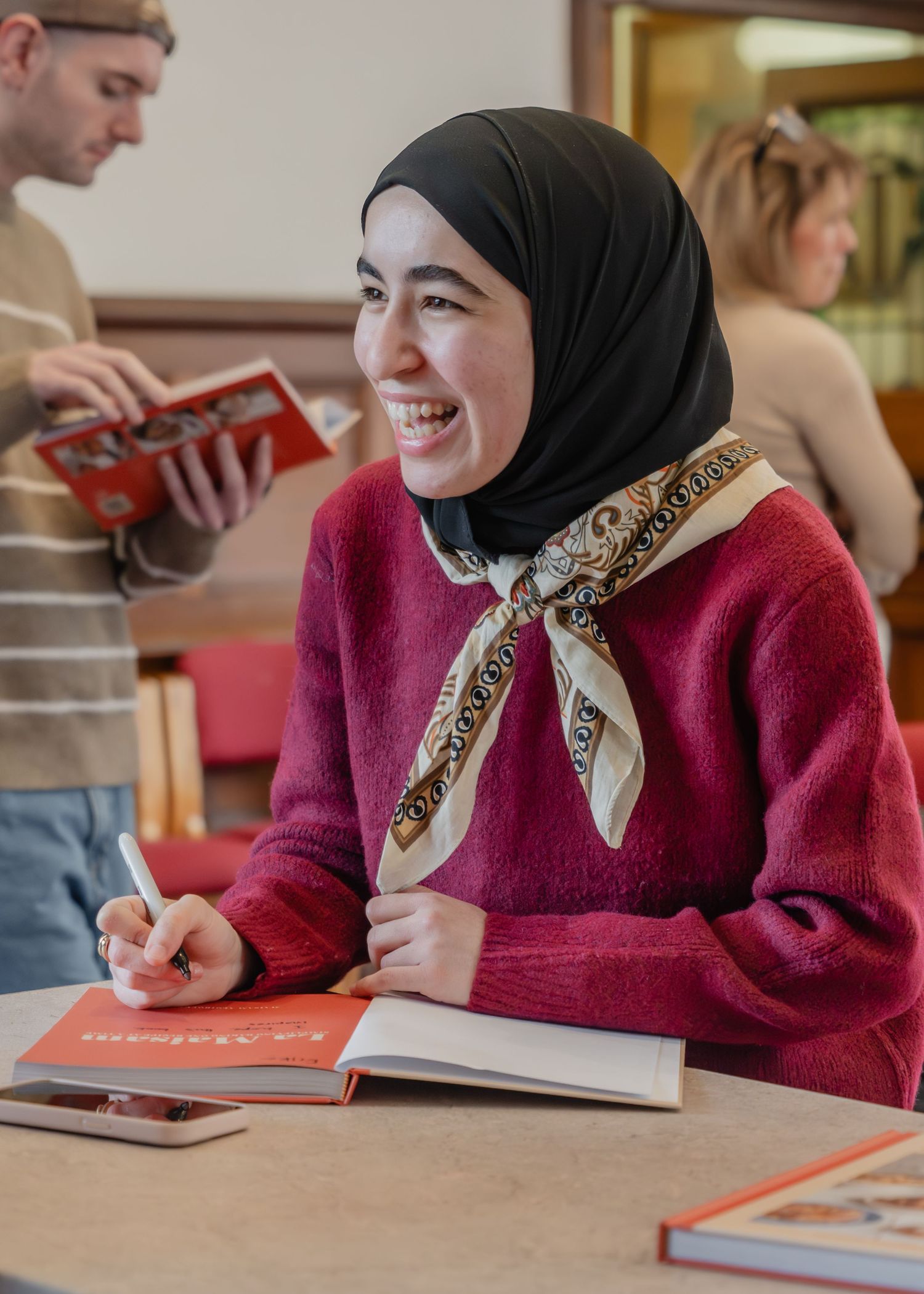 Student wearing a burgundy sweater and black hijab smiles while studying and taking notes in a classroom setting.