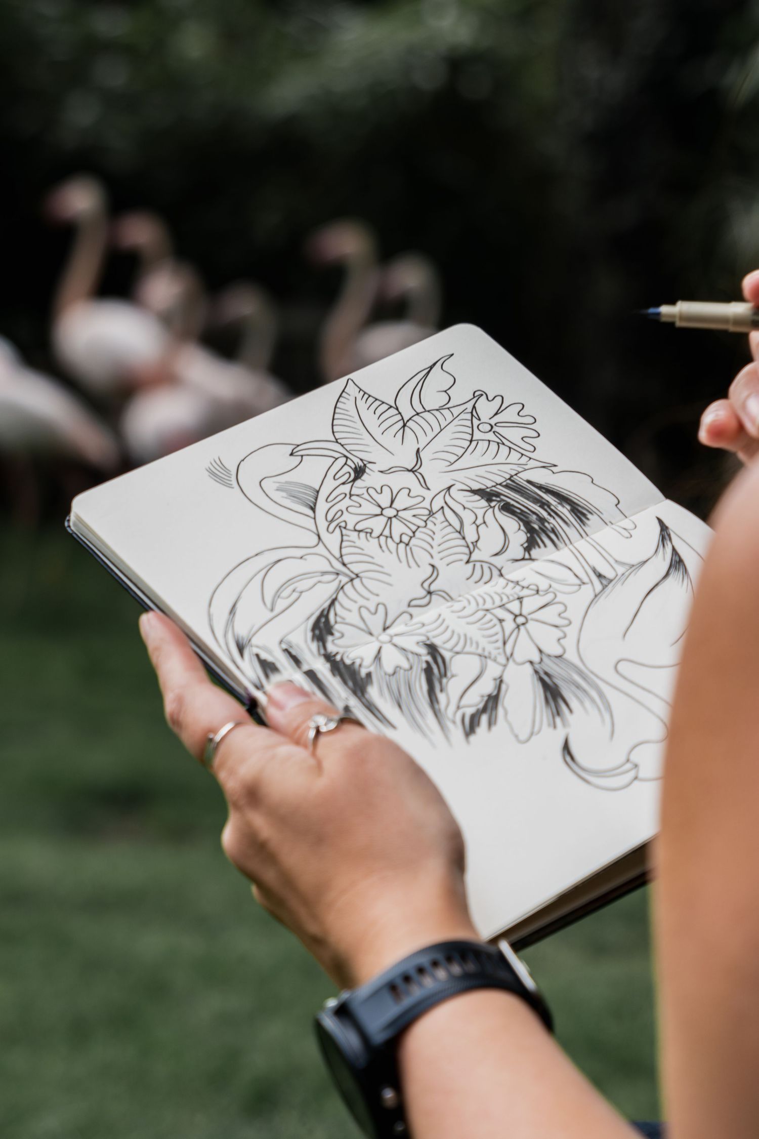 Artist sketches flowing organic designs in a notebook held against a natural outdoor background with blurred trees.