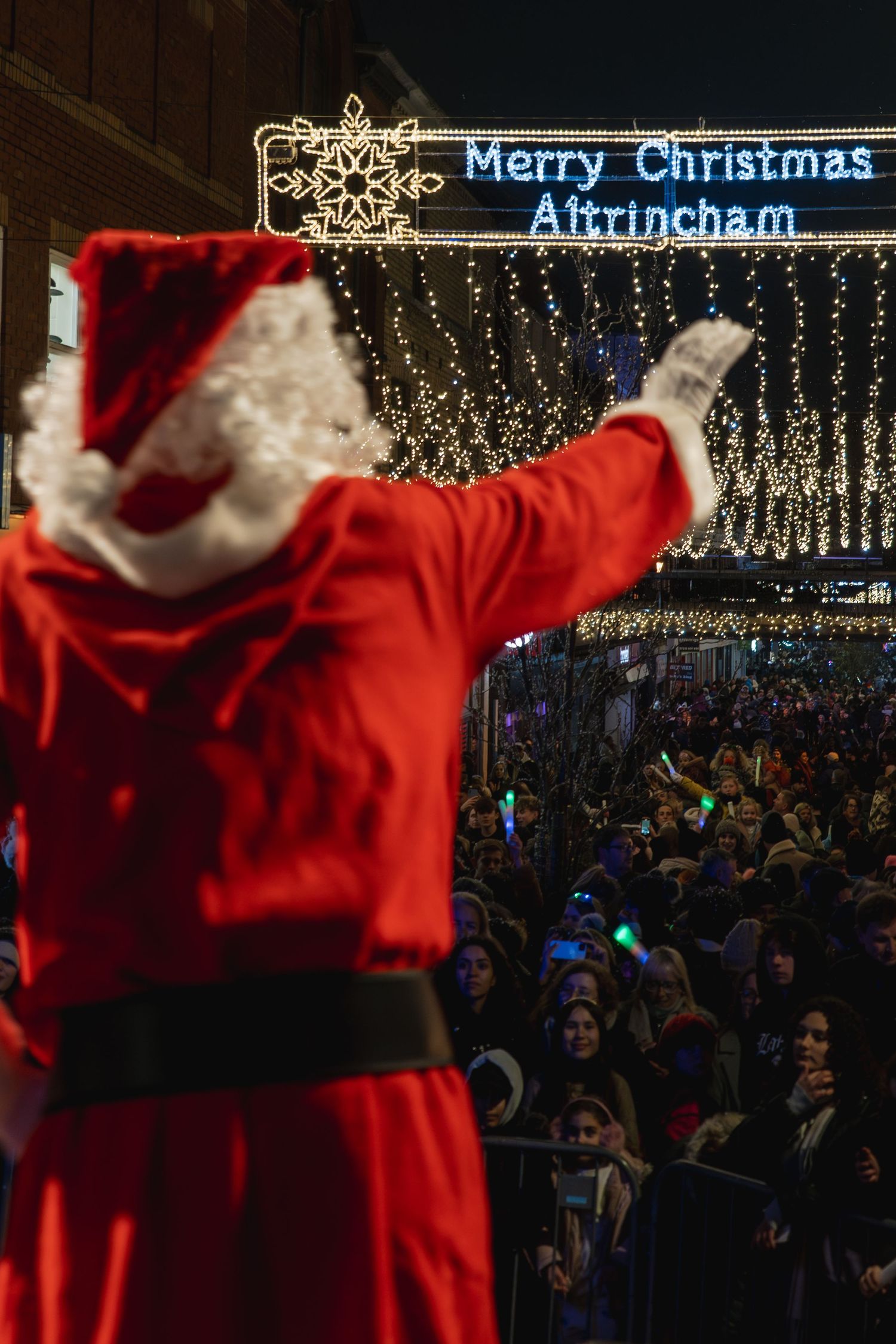 A festive crowd gathers under illuminated Christmas lights and decorations at a nighttime holiday celebration in Altrincham.