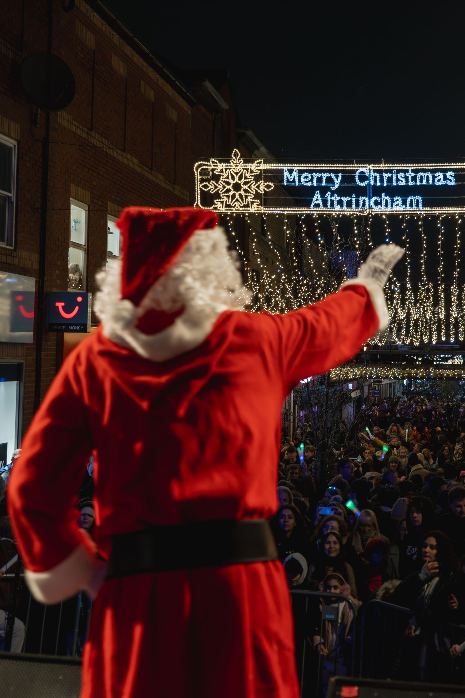 A figure in a red suit gestures beneath illuminated 'Merry Christmas Altrincham' sign at a nighttime holiday celebration.