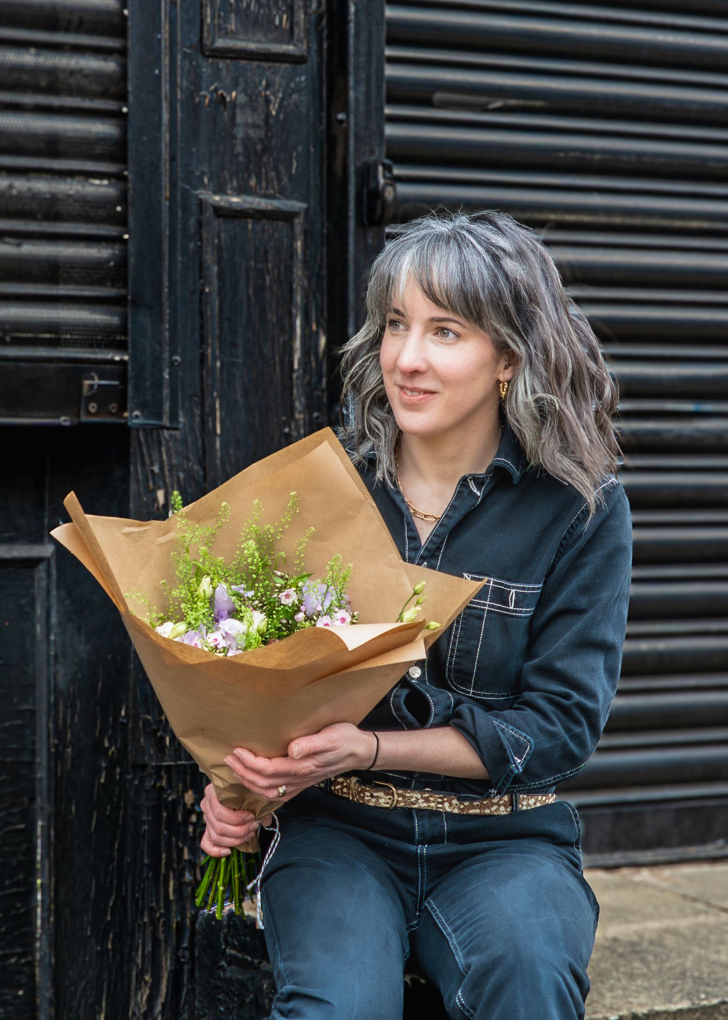 A person in denim clothing holds a fresh bouquet wrapped in brown paper while sitting against an industrial door.