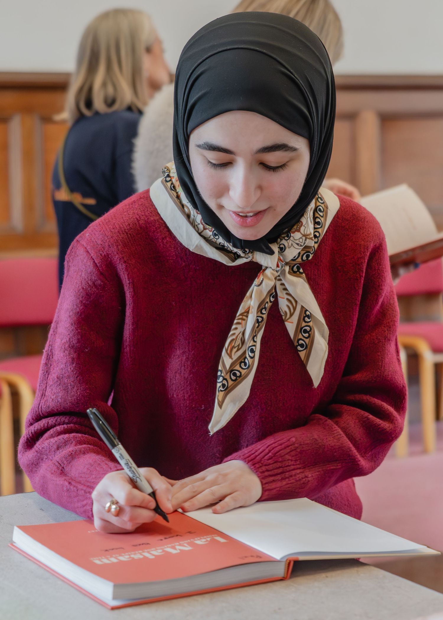 Student in a burgundy sweater and hijab writes in a book at a wooden desk in a library setting.