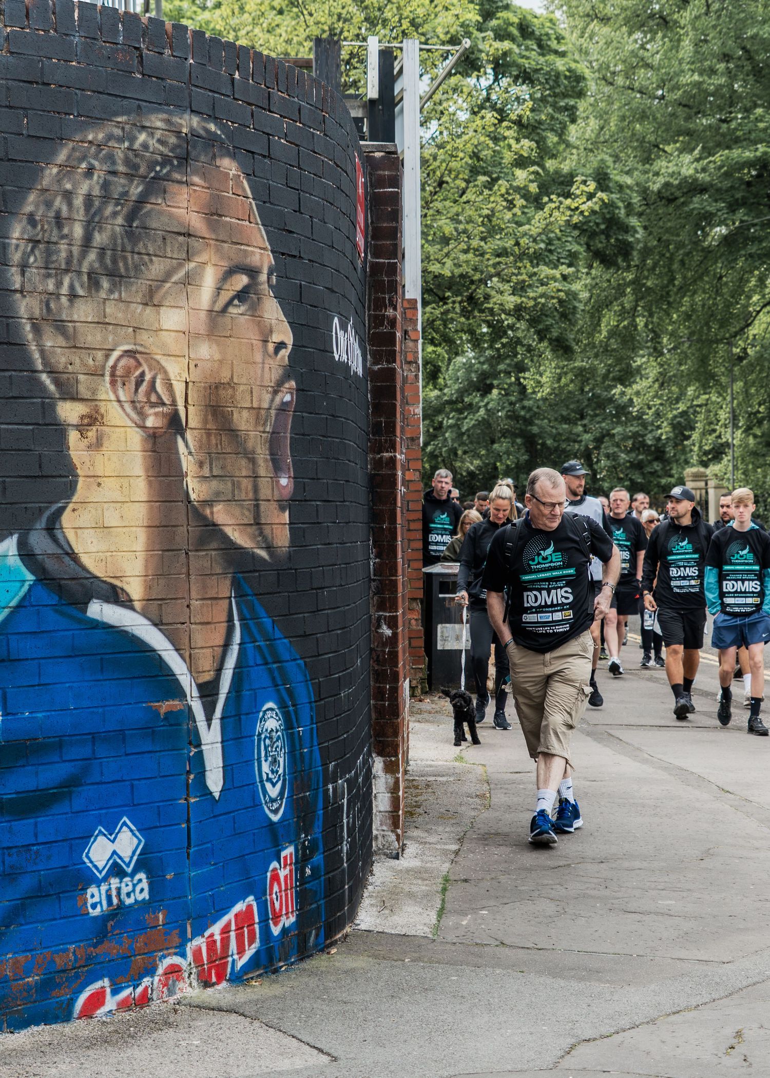 Group of people in matching black shirts walking past a large street mural of a footballer on a brick wall.