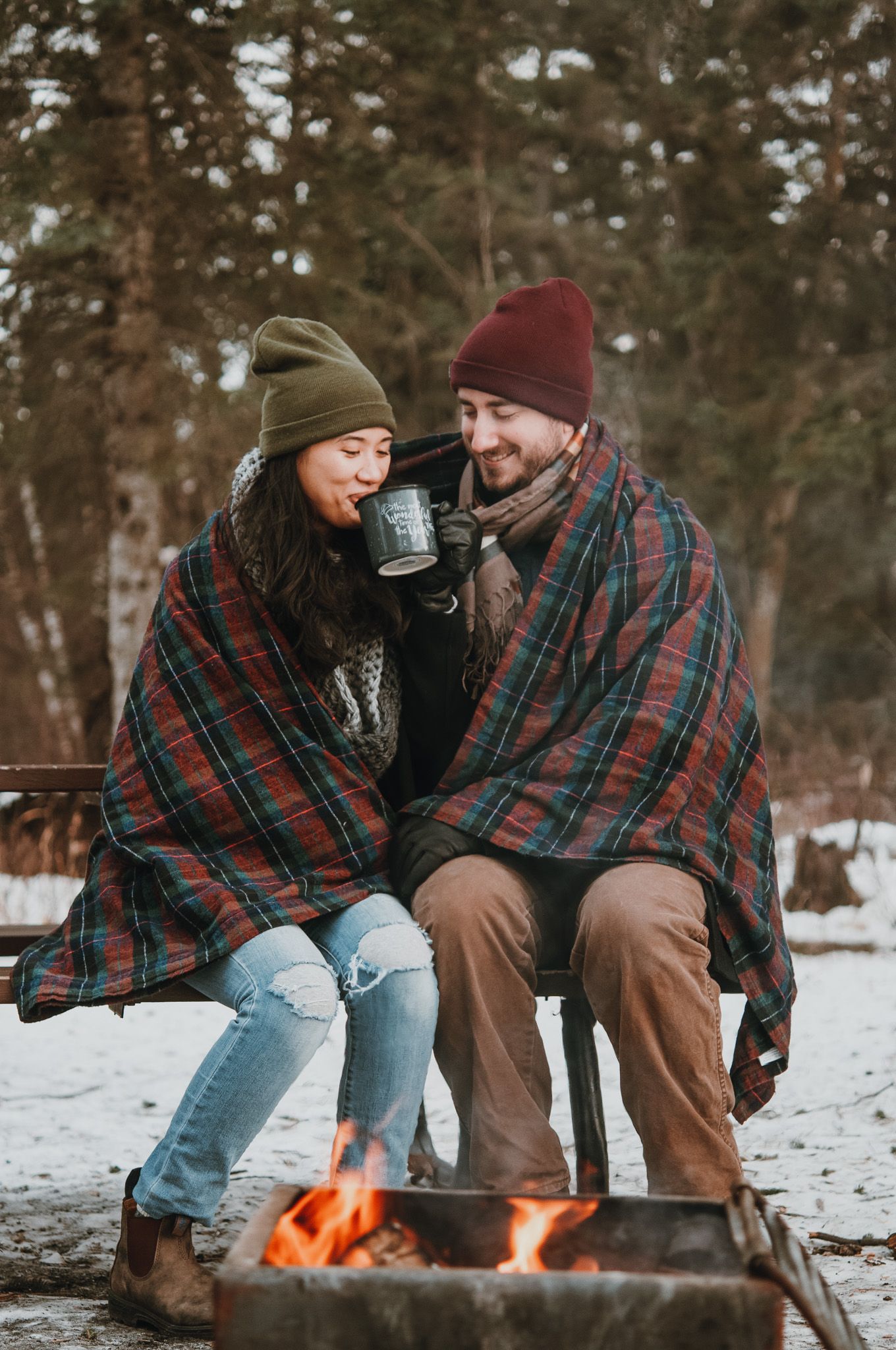 Couple sips coffee besdie fire outside next to fire, in winter forerst.