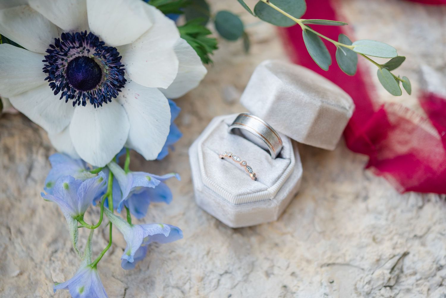 a couples wedding ring sit in a beige velvet ring box on a limestone rock, with the brides bouquet.