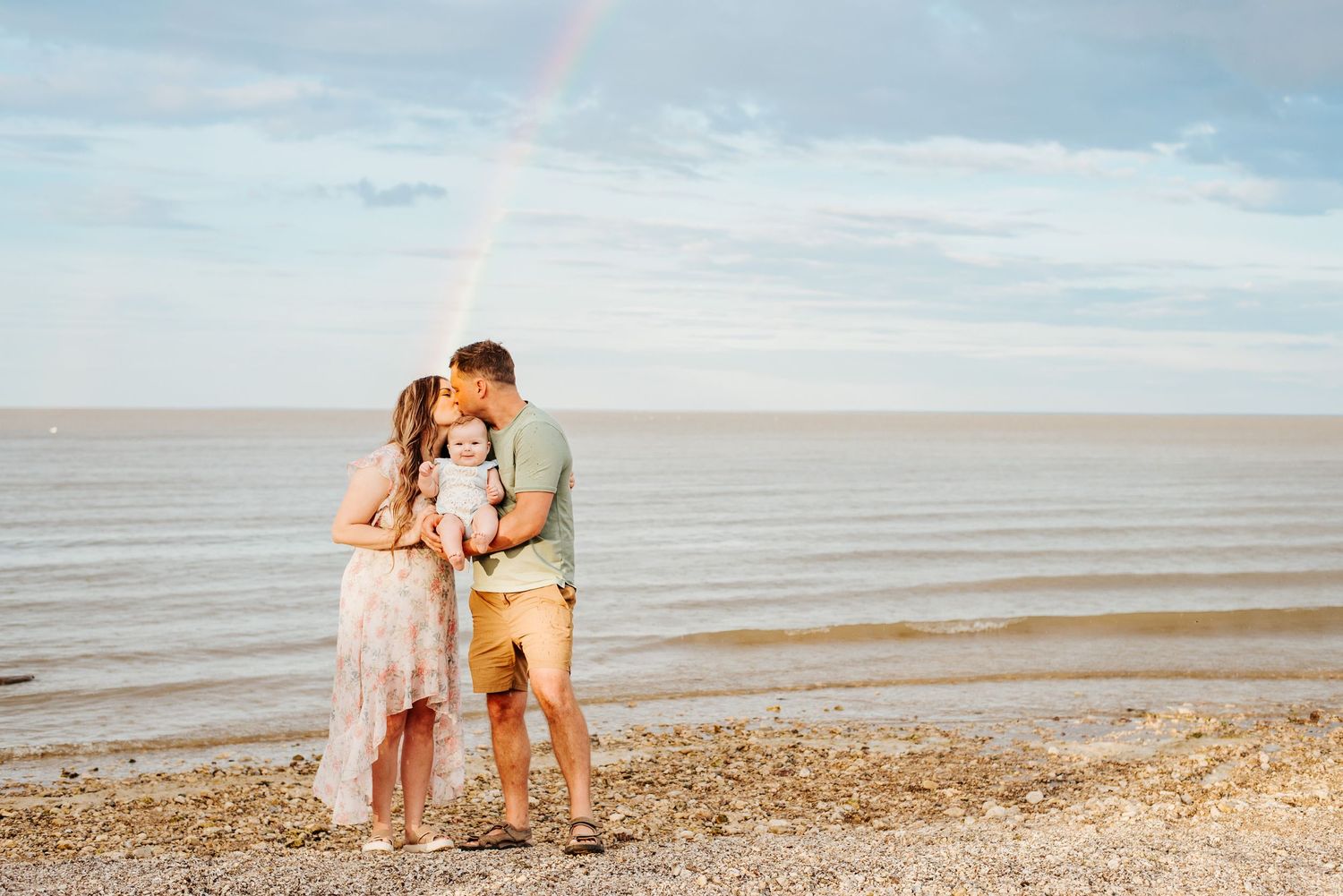 Mom and dad kiss holding baby with rainbow in the sky at the beach