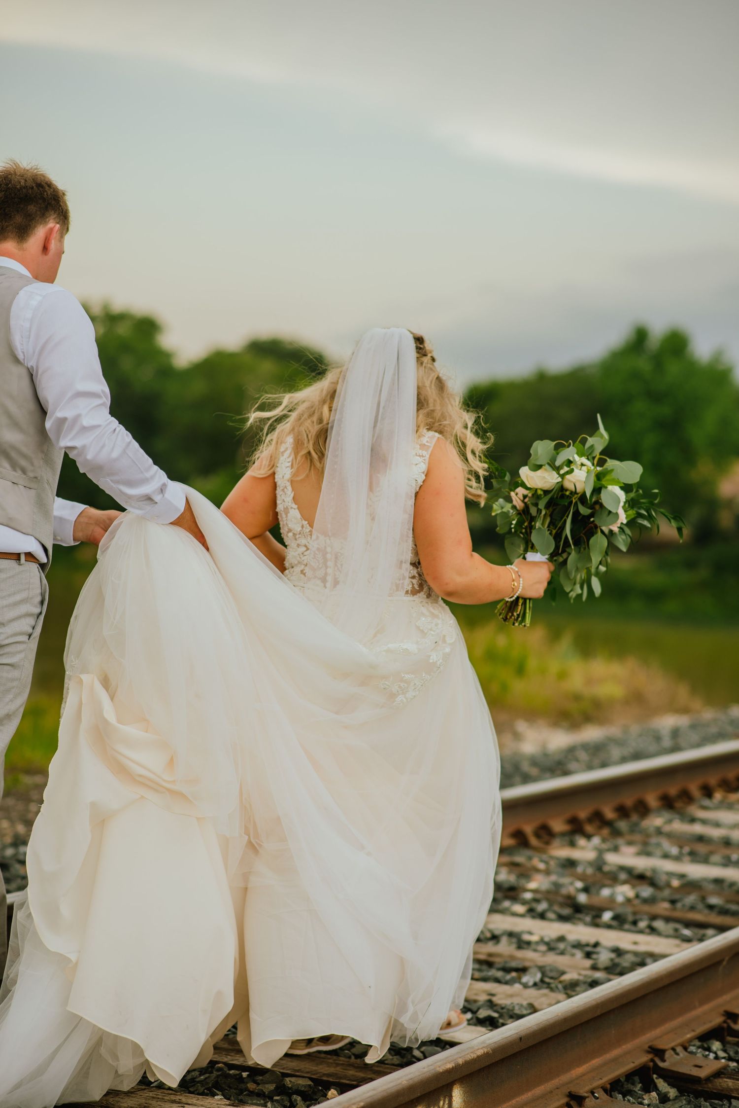 Man holds brides dress walking down railway