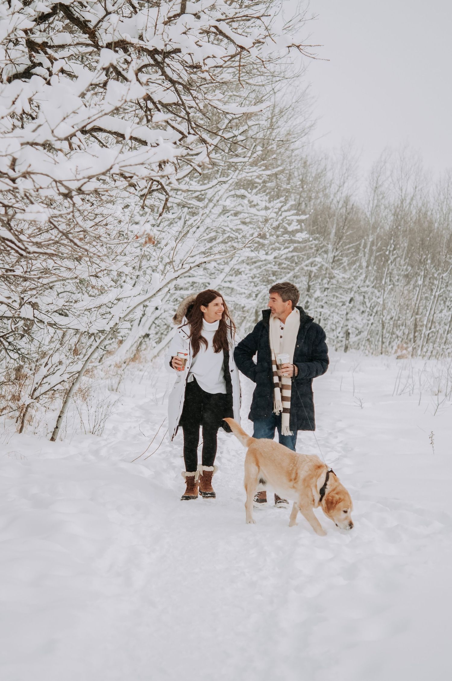 Couple walks dog while sipping coffee in a snowy winter wonderland for a professional photoshoot.
