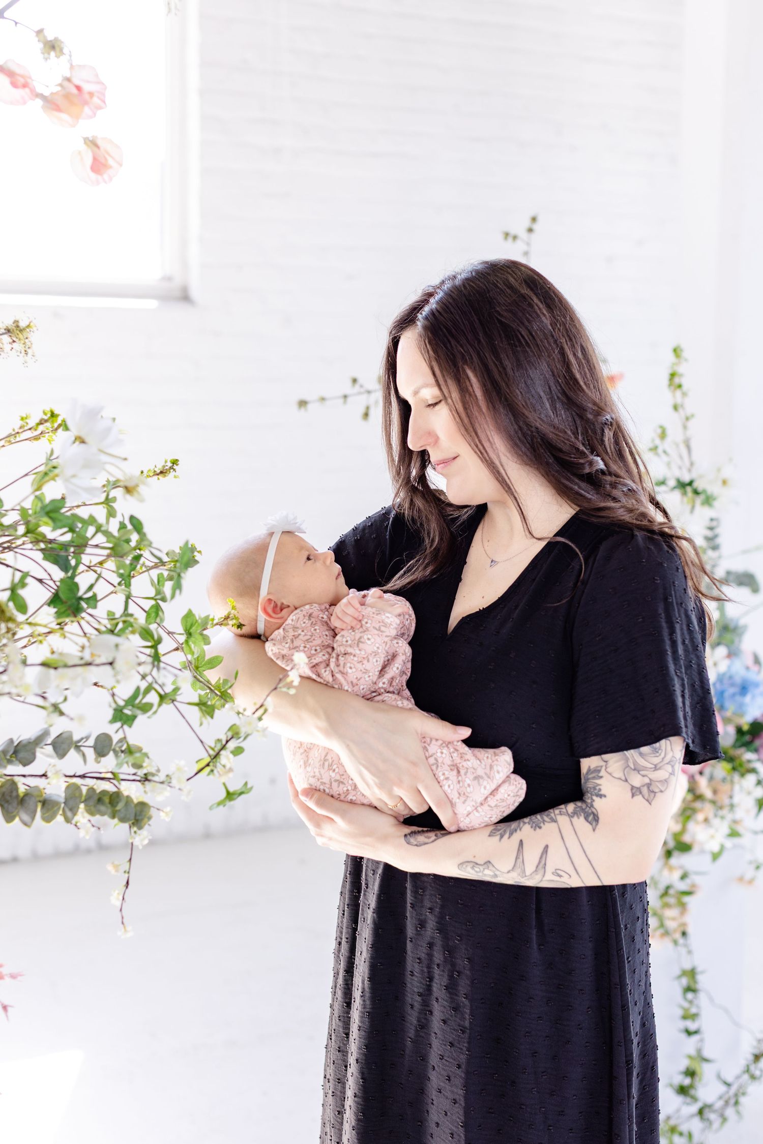 Mother wearing black dress with ling dark brown hair holding baby in pink clothes, in professional photo studio in Winnipeg for mothers  day