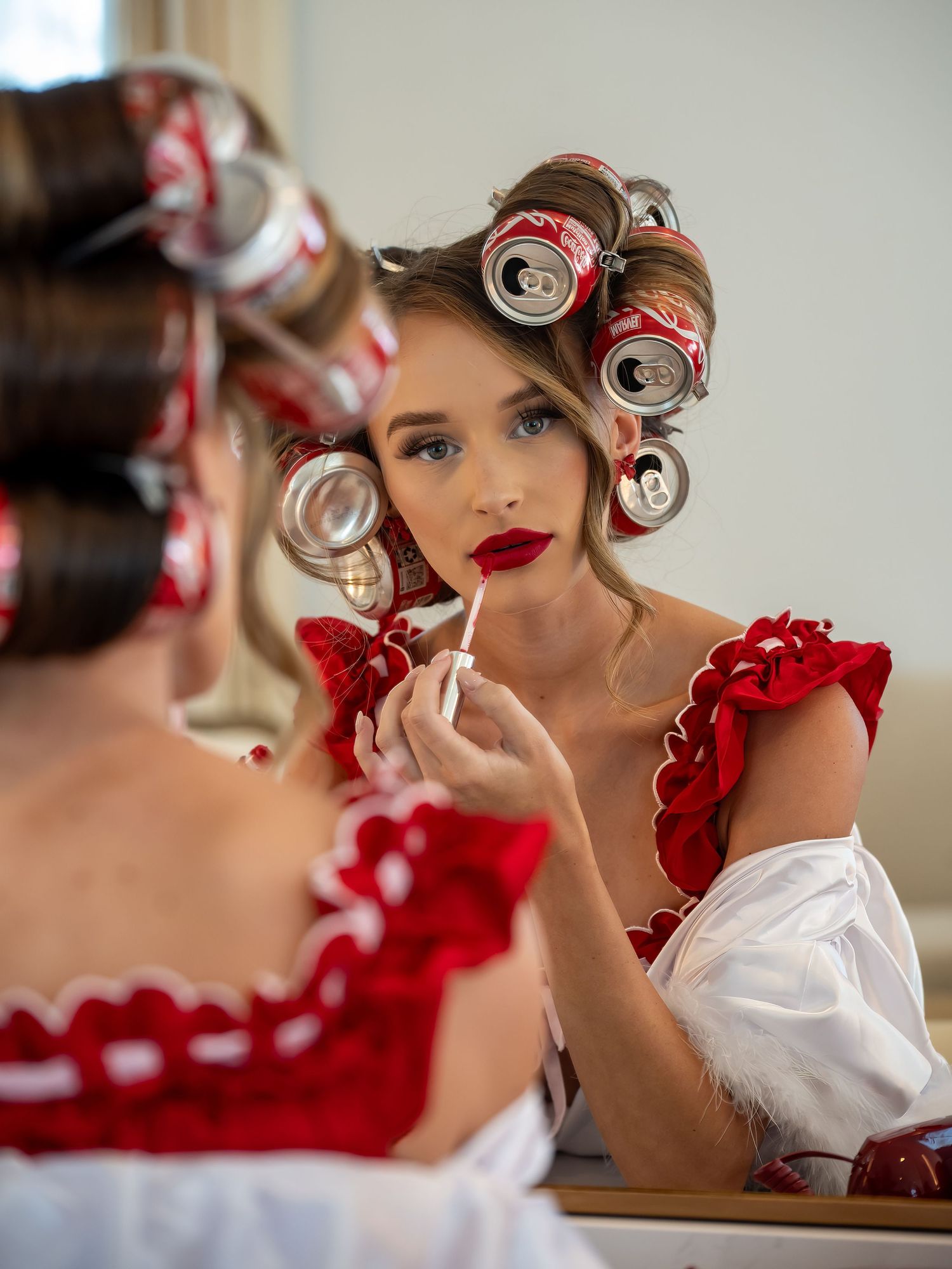 Woman applying red lipstick in mirror, wearing red ruffled dress with soda cans as hair rollers.