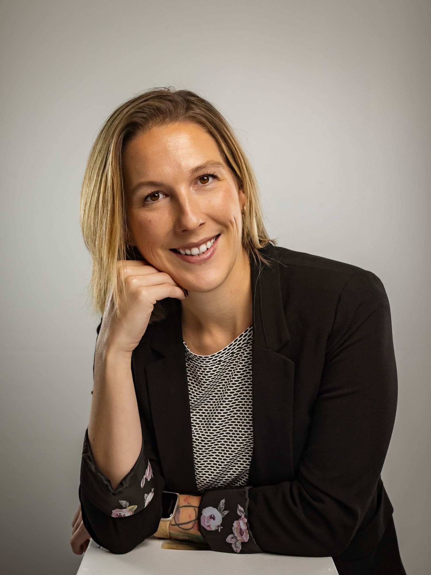 Smiling professional woman in black blazer resting chin on hand in relaxed business portrait.