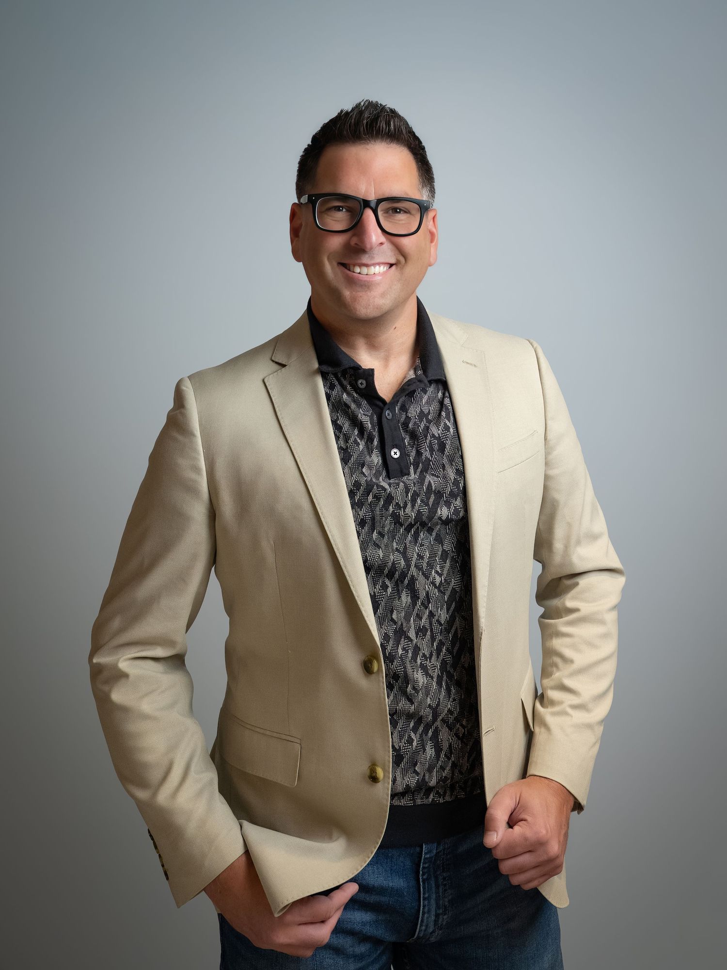 Smiling man in beige blazer and patterned shirt wearing glasses poses confidently for a professional portrait photo.