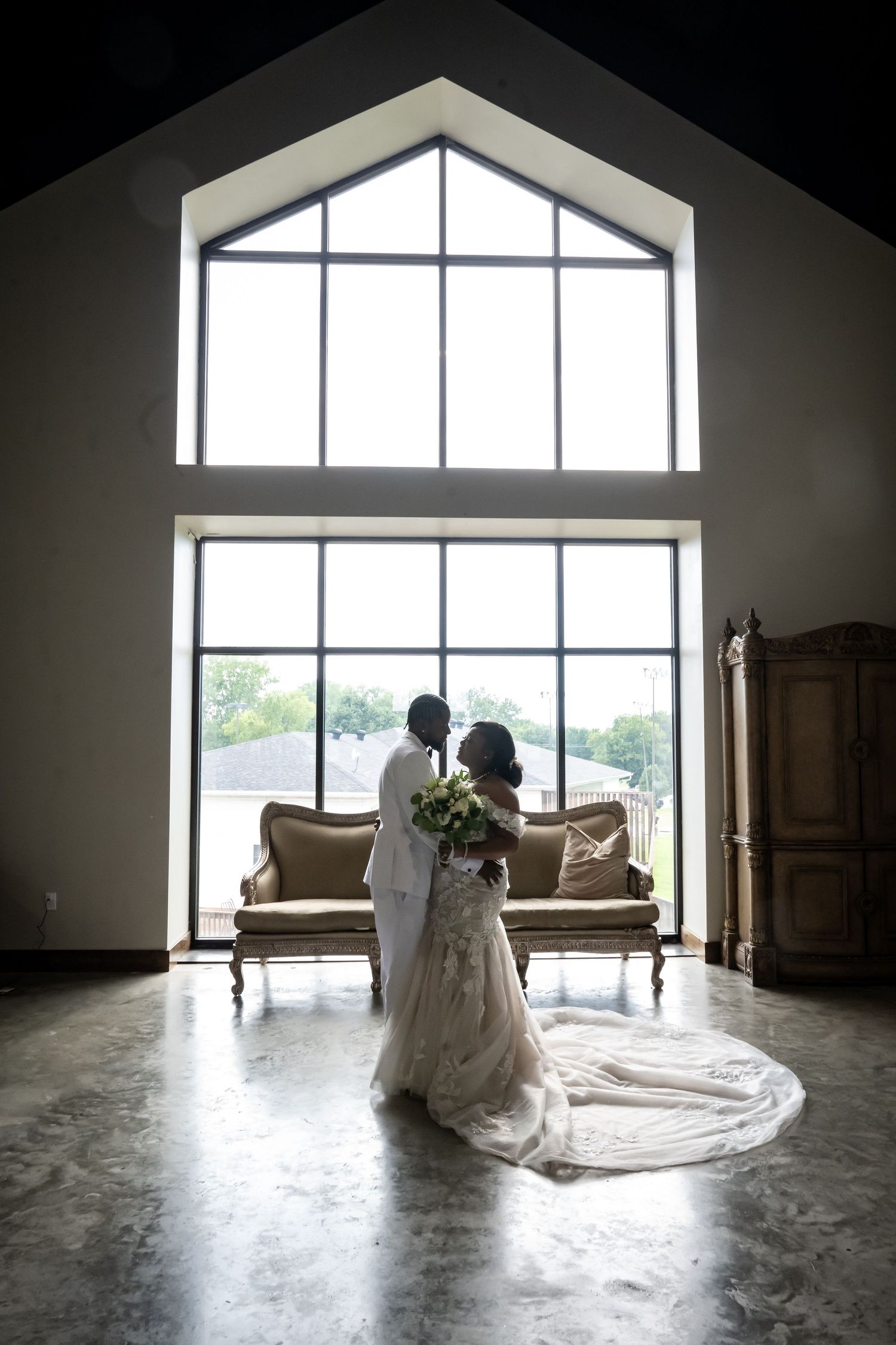 A bride and groom share an intimate moment in front of large windows in an elegant room.