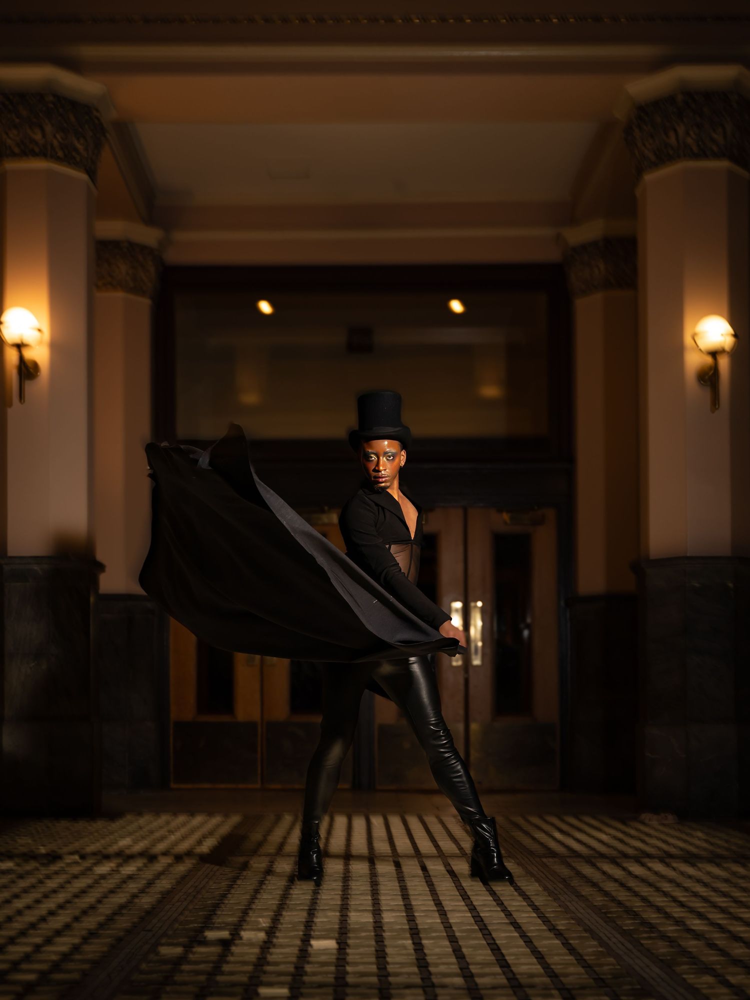 Dramatic silhouette in flowing black dress and hat poses in historic hallway with columns.