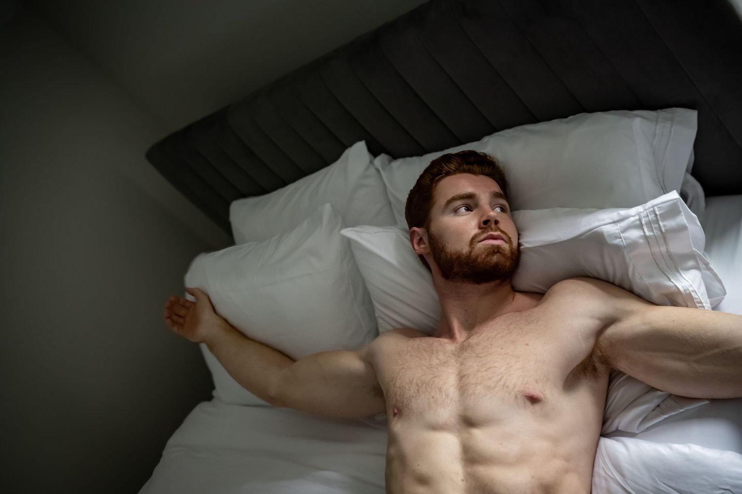 Shirtless redhead man with beard lying in bed on white pillows, looking upward in a dimly lit bedroom.