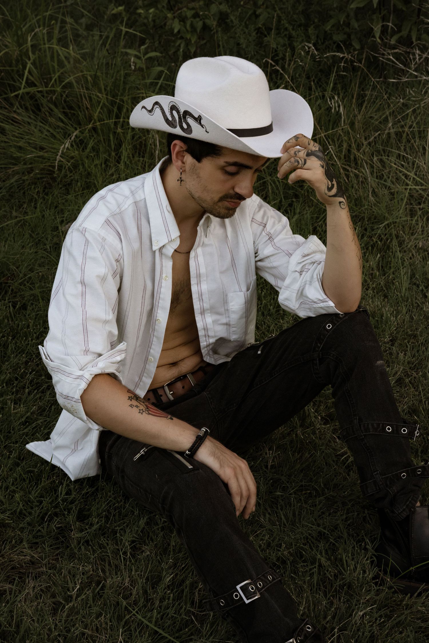 Young man in white cowboy hat and open striped shirt sitting on grass with tattoos and dark pants.
