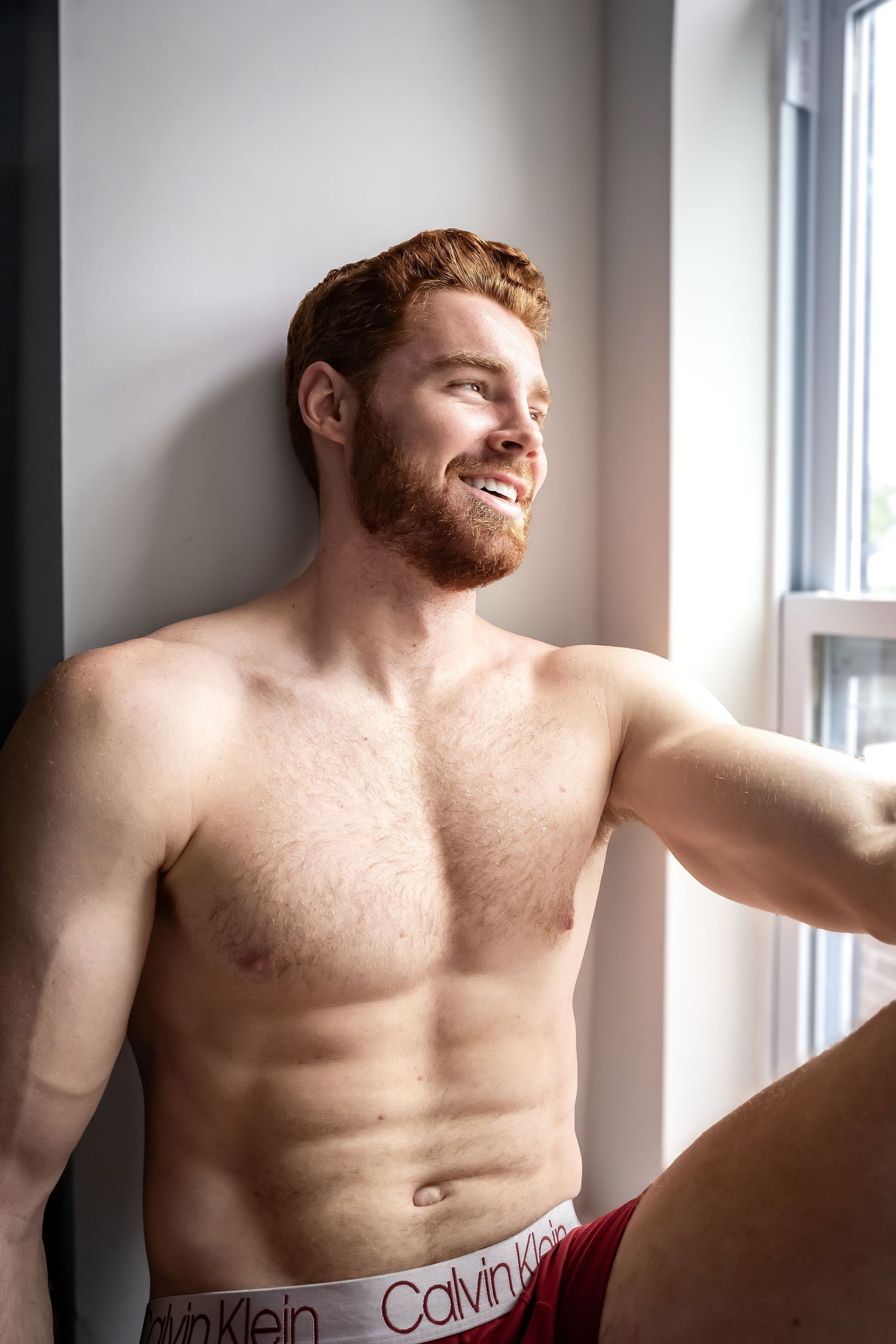 Athletic shirtless person in Calvin Klein underwear poses near window in bright natural lighting.